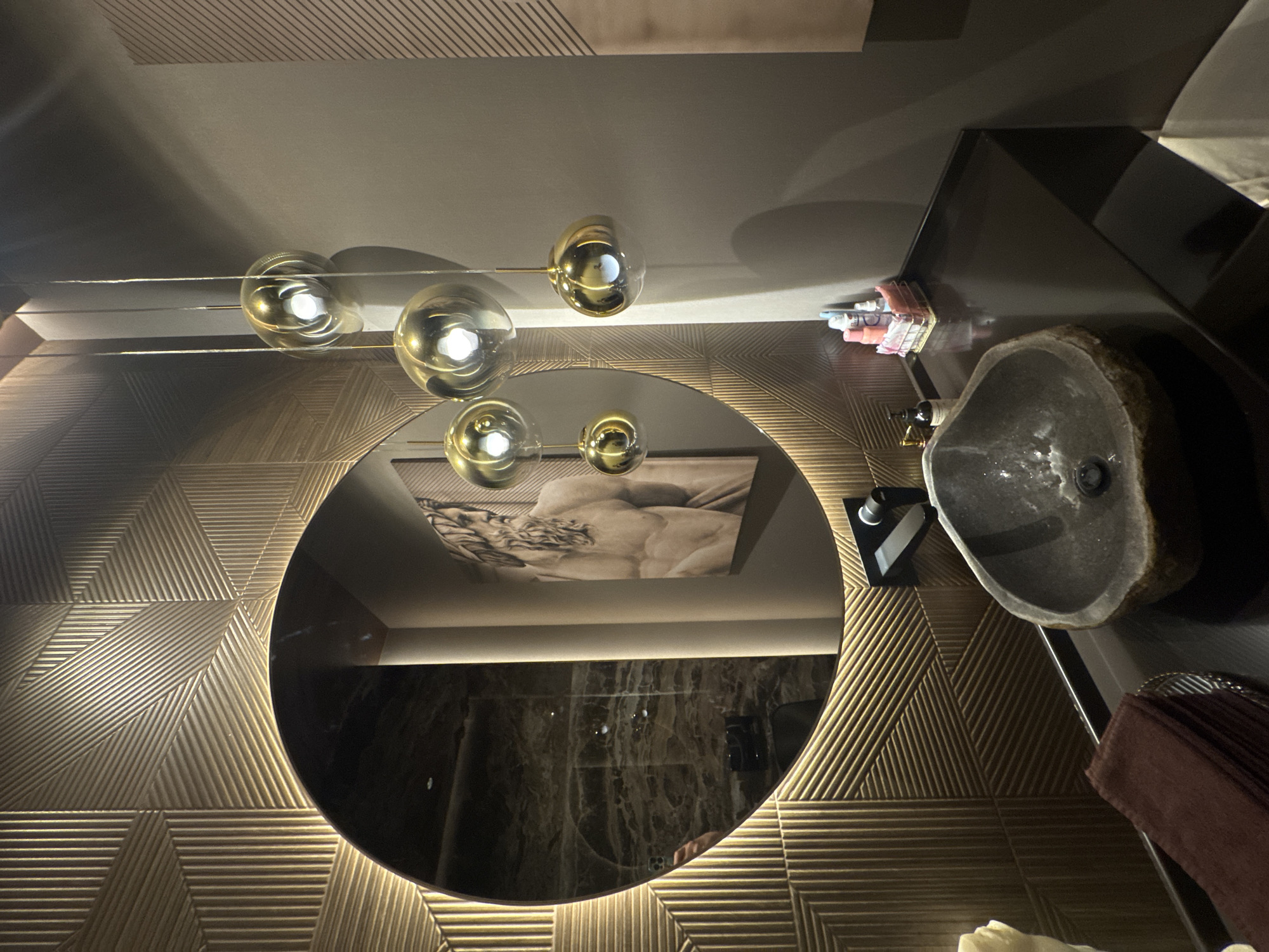 This is an indoor scene featuring a modern bathroom vanity area. A large, circular mirror with integrated lighting is mounted on a textured, gold-colored wall. To the right of the mirror, a painting of a muscular, classical male figure is displayed. Above the vanity, a cluster of pendant lights with spherical, golden-tinted glass shades hangs.
The vanity itself is made of a dark, reflective material. A unique, irregular-shaped sink, appearing to be carved from stone, is set into the vanity. A dark-colored faucet is mounted behind the sink. On the vanity countertop, to the right of the sink, are several small bottles of toiletries arranged in a wire basket. To the left of the sink, a stack of dark red towels is placed in a metallic basket.
The wall to the right of the vanity is painted a neutral gray and features a subtle striped texture. The overall aesthetic is luxurious and contemporary, with a blend of metallic finishes, natural textures, and artistic elements.