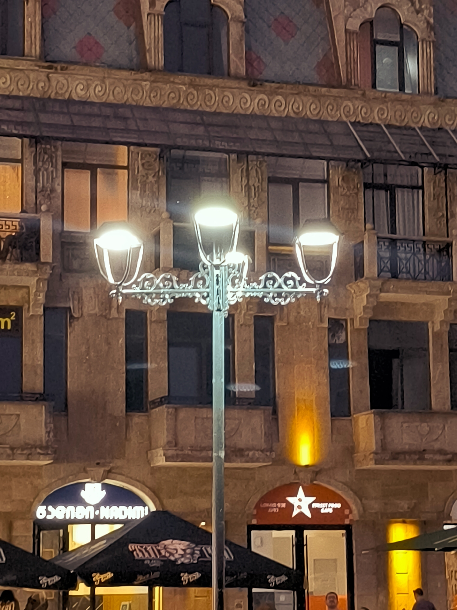 This is a night-time, eye-level shot of a street in Batumi, Georgia, likely taken near the intersection of Rustaveli Avenue and Ninoshvili Street, given the presence of the "STREET FOOD CAFE" and the architectural style. The scene is dominated by the illuminated facade of an old building with ornate architectural details, including decorative cornices and columns. A streetlamp with three lights stands prominently in the foreground, casting a warm glow. On the ground level, two establishments are visible: one with an arched entrance displaying a sign in Georgian script ("გონონ•NADIMI") and a dark umbrella in front, and another with an arched entrance featuring a white star logo and the words "STREET FOOD CAFE" on a red sign. Several umbrellas, likely for outdoor seating, are visible. Two individuals, a man and a person whose face is not clearly visible, are present in the lower right portion of the image, appearing to be walking or standing. The overall impression is that of a lively evening street scene in a historic urban area.