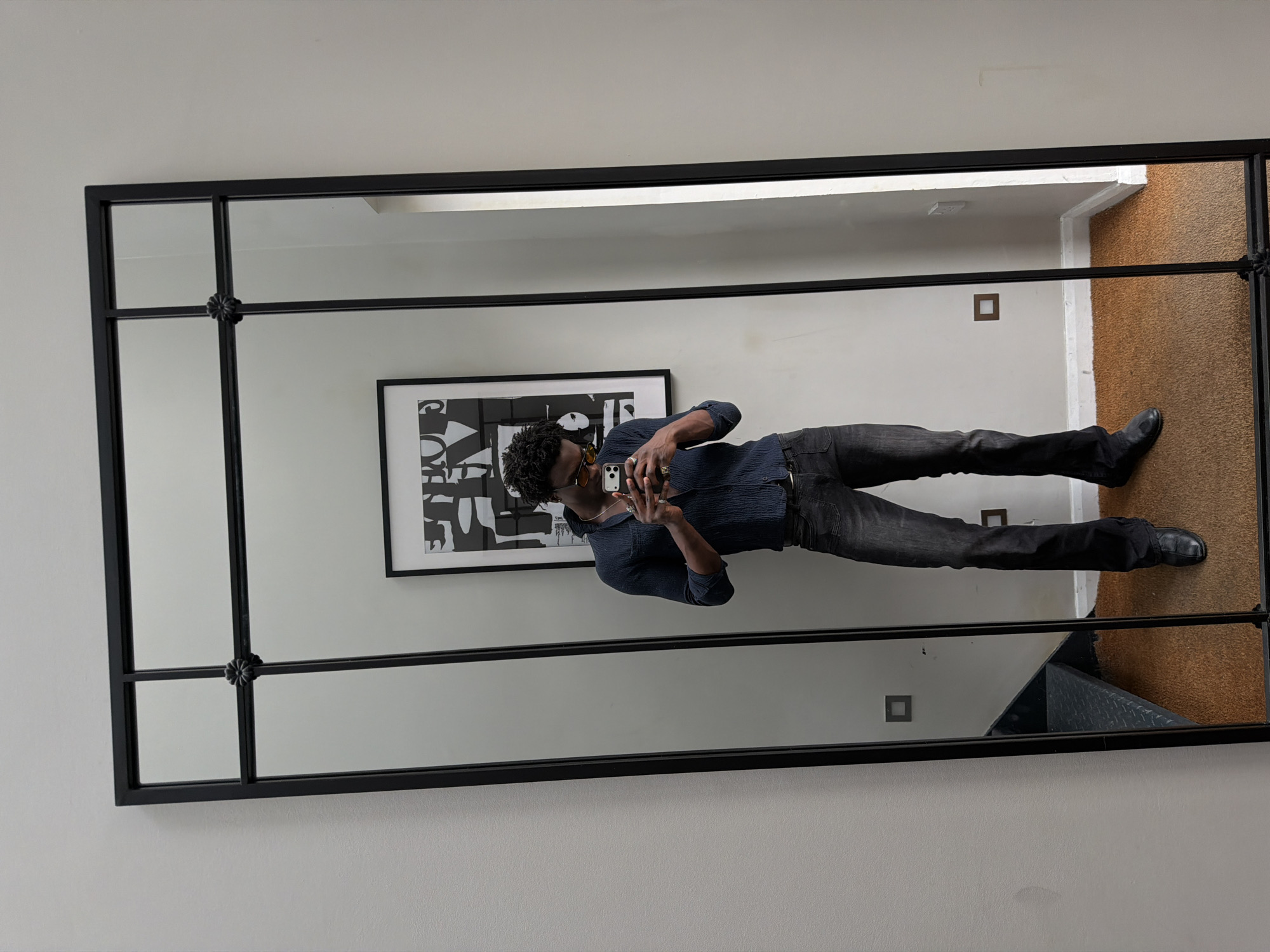 The image displays a full-length mirror selfie of a person, rotated 90 degrees counter-clockwise. The central subject is a person, likely male, with dark, curly hair, dressed in a dark blue long-sleeved shirt, dark jeans, and black shoes. They are holding a white smartphone (appearing to be an iPhone) in front of their face to capture the reflection. The person is wearing sunglasses or tinted glasses, a ring on their right hand, and a necklace.
The large mirror features a prominent black metal frame, divided into multiple rectangular sections by thinner black metal bars. A decorative, black floral-like element is fixed at the intersection of the horizontal and vertical bars on the left side of the frame.
The setting reflected in the mirror is an indoor space with light-colored walls (off-white or light grey). Behind the person, a framed black and white abstract or typographic art piece is visible on the wall. The floor in the reflection shows a brown carpeted area on the right, and a lighter floor, possibly wood or tile, on the left. A glimpse of dark grey metal stairs is also visible in the lower right reflection. Small, square wall fixtures are present on the reflected wall. The lighting appears bright and even, consistent with an indoor environment. No readable text is visible in the image. The location context is London, United Kingdom.