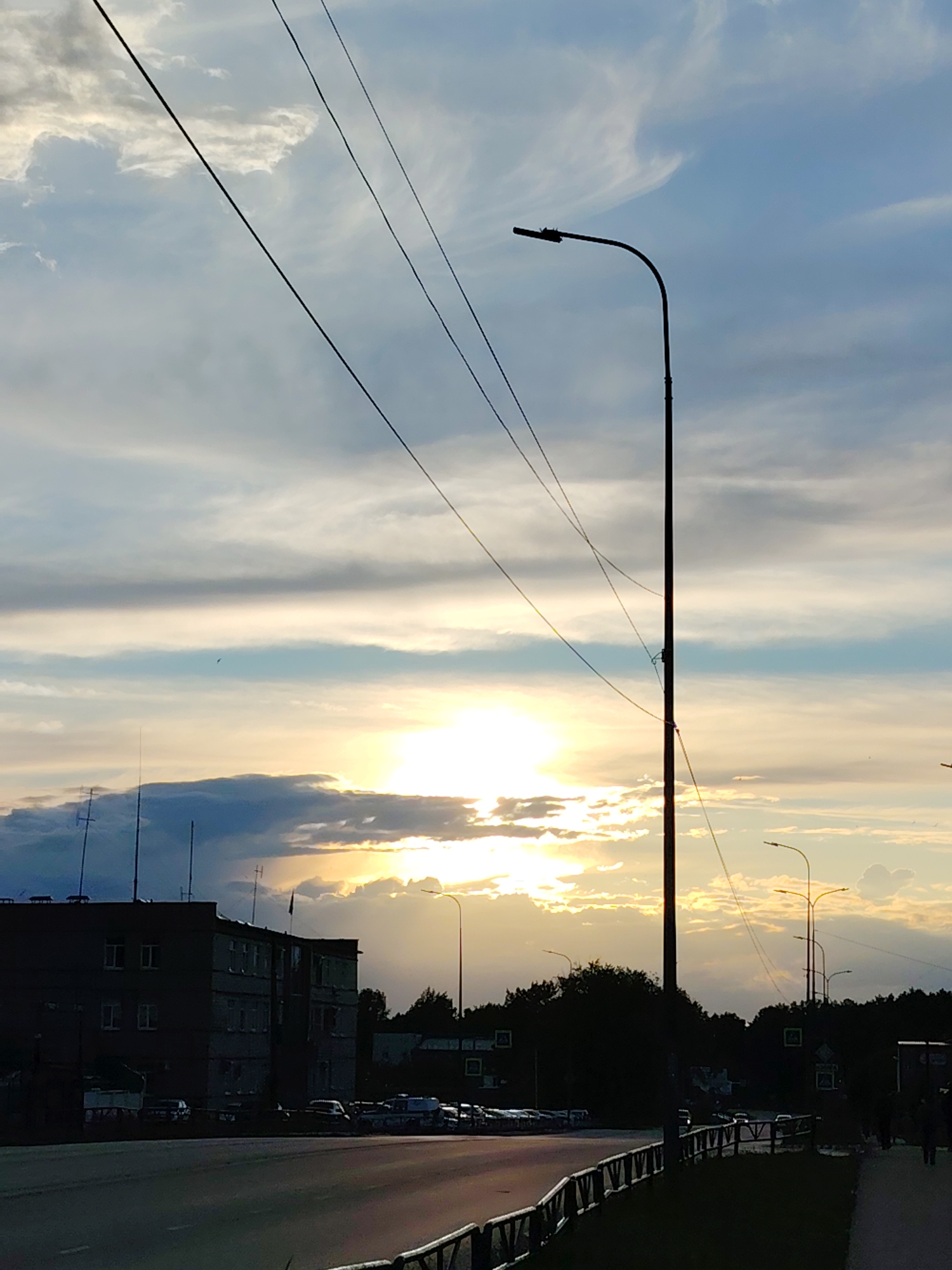 The image captures a street scene in Chaikovsky, Russia, during a sunset or sunrise. A multi-story building is situated on the left side of the frame, with antennae visible on the roof. A line of parked cars is visible in front of the building. A road runs across the foreground of the image, separated from a grassy area and sidewalk by a guardrail. A tall street light is located on the right side of the frame, with other street lights visible in the distance. Power lines stretch across the sky. The sky is filled with clouds and the sun is visible through a break in the clouds. A few indistinct figures are present on the sidewalk on the right side. The overall lighting suggests either early morning or late evening.