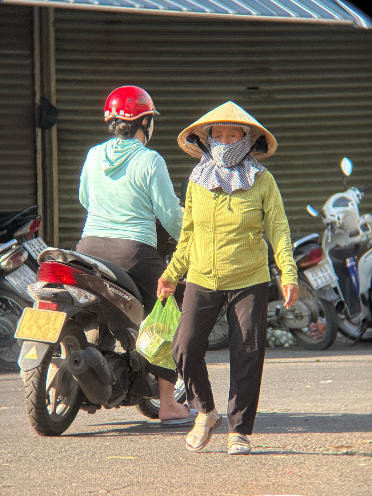 Two individuals are on motorcycles in a city setting, likely Nha Trang, Vietnam. The person in the foreground is wearing a conical hat, a checkered scarf covering their face and neck, and a light green long-sleeved shirt with black pants and sandals. They are carrying a translucent green plastic bag in their right hand. The motorcycle in front of them is dark brown with a yellow license plate and a visible "VISION" logo. The second person, partially visible on the left, is wearing a red helmet and a light blue hooded jacket, also seated on a motorcycle. The background features a corrugated metal shutter and several other motorcycles parked nearby. The ground is asphalt, with shadows indicating it is daytime.
