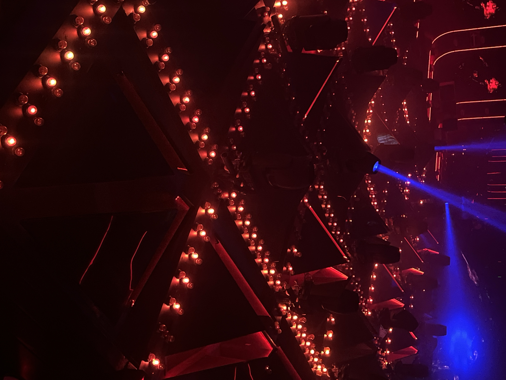 The image shows an interior space with a dark, club-like atmosphere. Triangular structures decorated with numerous small, warm-toned lights dominate the scene, appearing to create a geometric pattern or wall. A blue light beam cuts through the right side of the image. The location is Unknown, Unknown.