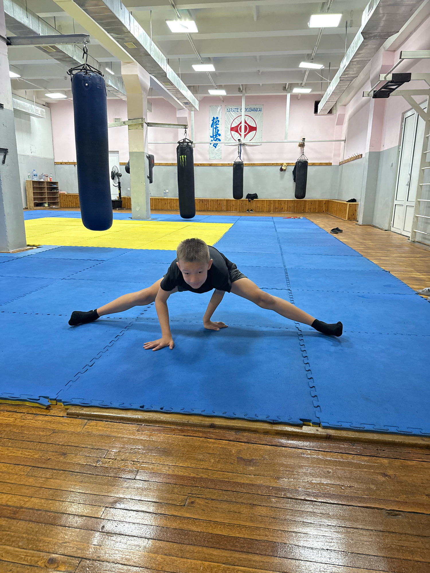 A young boy with short brown hair, wearing a black t-shirt and black socks, is performing a split on a blue mat in a gymnasium. His body is angled towards the camera, with his hands on the mat for support. His legs are extended wide apart, with his left leg forward and his right leg back. The gymnasium has a wooden floor and a yellow mat in the background. Several punching bags hang from the ceiling, and a banner with Japanese characters is displayed on the wall. The lighting suggests an indoor setting. The scene depicts a training or practice session, likely for a martial art or physical conditioning.