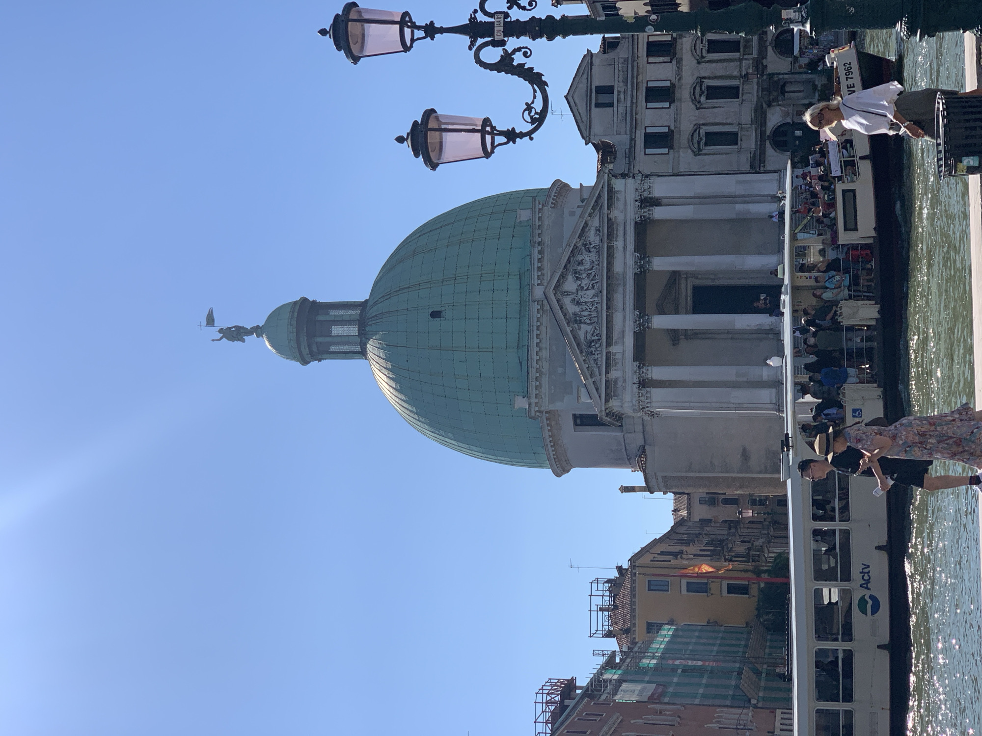 The image depicts a sunny day in Venice, Italy, with a prominent domed building dominating the skyline. This building, likely the San Simeone Piccolo church due to its characteristic green dome and neoclassical facade, is situated on the Grand Canal. In the foreground, a water taxi, identified by the "Actv" logo, is visible. Several tourists are boarding or disembarking from the water taxi. On the left side of the image, a couple walks along the canal's edge, while on the right, a woman stands near a trash receptacle. The canal is shimmering with sunlight, reflecting the bright blue sky. The overall scene captures a typical moment of urban life and tourism in Venice.