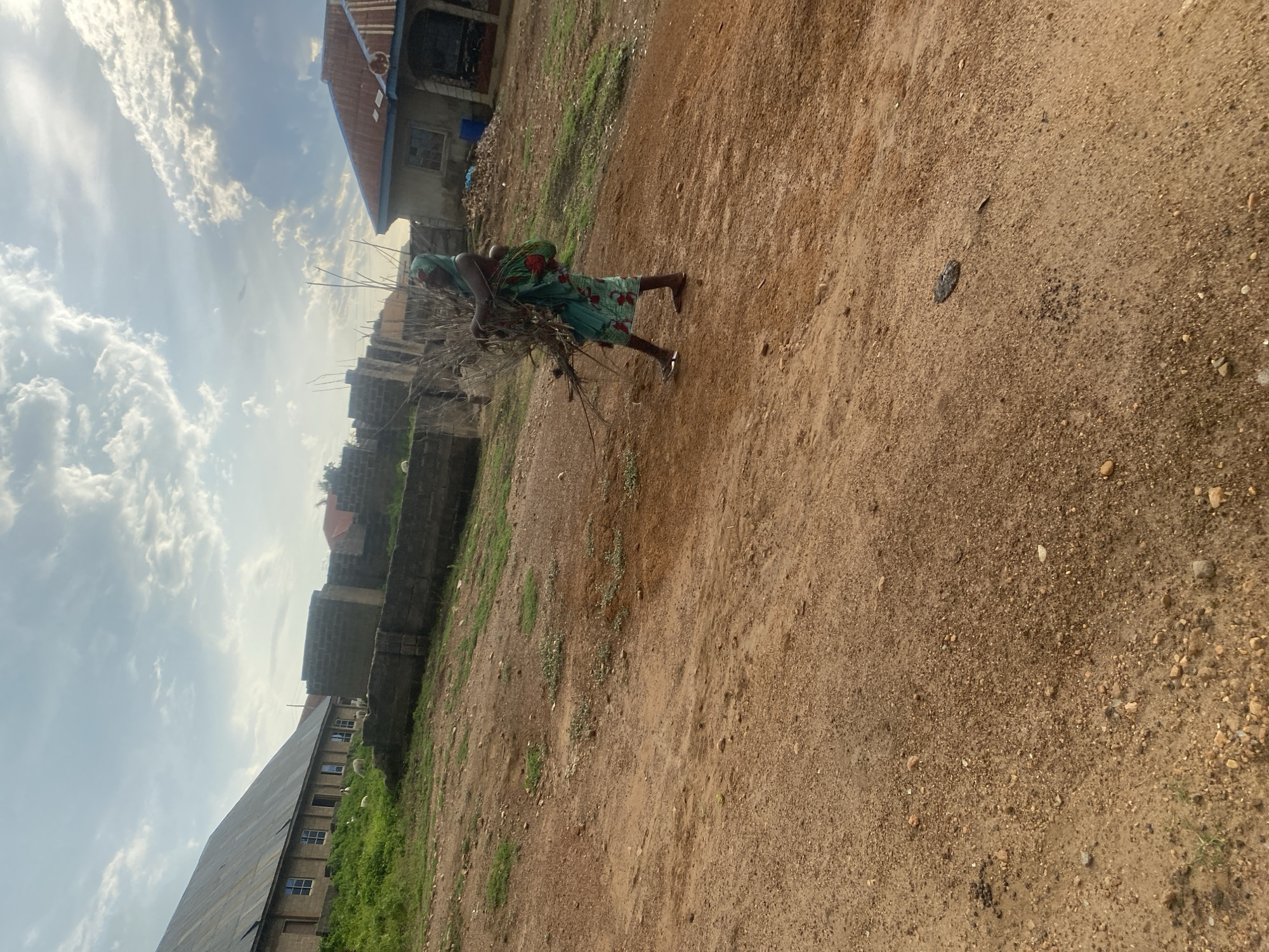 In an outdoor, dusty, and rural environment, a person wearing a headscarf and a patterned dress is carrying a bundle of sticks on their back, with a child visible on their back as well. The person is walking on a dirt path. In the background, there are several buildings with corrugated metal roofs and brick walls, some of which appear to be under construction. There is also some sparse green vegetation and a cloudy sky. The scene suggests a simple, possibly impoverished, living condition.