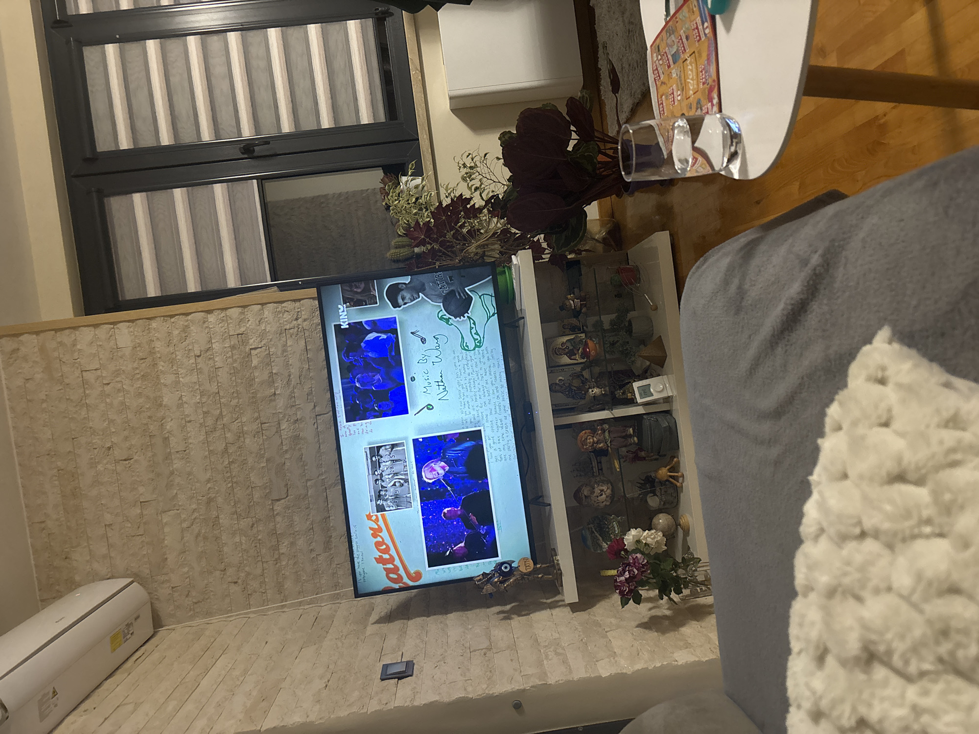 This image captures an indoor living room in Blagoevgrad, Bulgaria, from a low, tilted perspective, likely taken from a sofa. The scene is dominated by a flat-screen television displaying a multimedia montage, possibly a music video or tribute. The TV shows multiple frames, including images of individuals and groups, set against blue-toned backgrounds. Text visible on the screen includes "Music By Nathan Warg" and various snippets like "the biggest smile of everyone I know!", "begin well our Senior year has", "I love you meling my core value", and the word "GATORS". Below the TV, a white air conditioning unit sits on the floor.
To the left of the TV, a large window with black frames is covered by striped grey and white roller blinds. The wall beneath and around the TV is clad with light-colored, stacked stone. On the right, a grey sofa with a fluffy white blanket is partially visible in the foreground. A small, round white side table holds two clear glasses. Behind the TV, a white shelf unit contains various decorative items, including figurines, a small clock, and a vase with white and purple/red flowers. Several potted plants with both dark and lighter foliage are placed on top and around this unit, adding greenery to the space. The floor visible near the table is made of wood. The lighting suggests it is evening or night indoors, with artificial light illuminating the room.