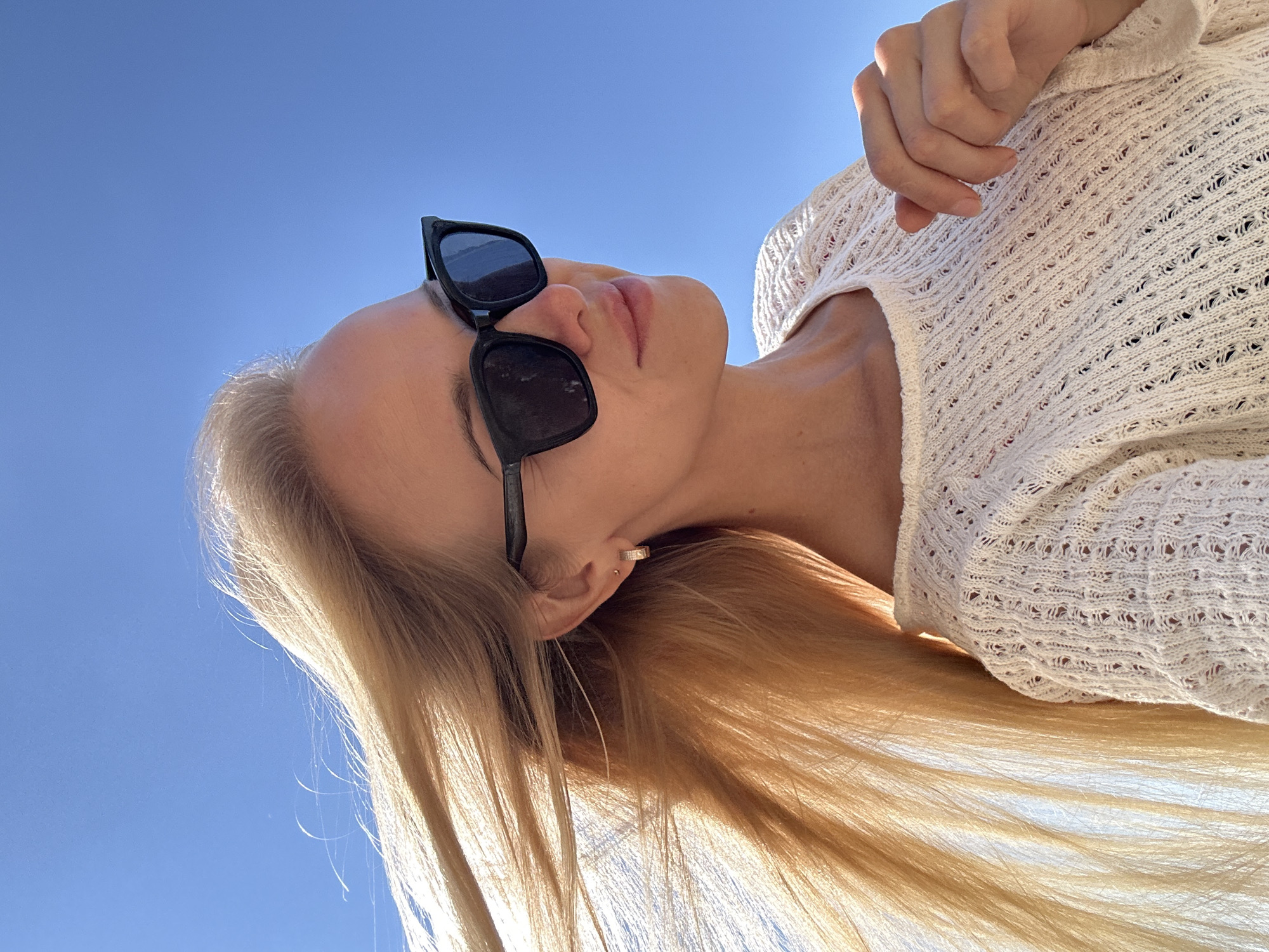 A low-angle, medium shot captures a blonde woman in sunglasses and a white knit sweater against a clear blue sky. Her long, blonde hair is blown by the wind, creating a dynamic visual. The woman's face is turned to her right, with a slight smile. She wears a small hoop earring in her visible ear and her right hand rests on her chest. The background is entirely the bright blue sky, suggesting an outdoor setting. The image does not depict the city of Gornyi Erik, Russia, or any urban environment.