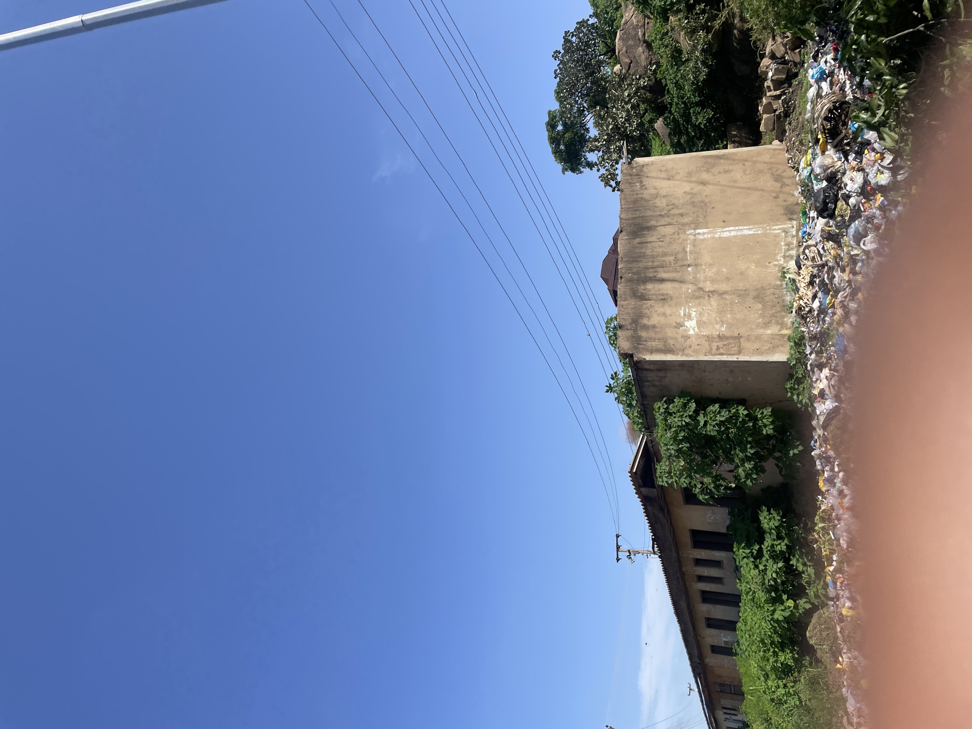 The image depicts a scene of urban decay in Jos, Nigeria. In the foreground, a large pile of uncollected garbage is visible, consisting of plastic bags, bottles, and other refuse. Behind the trash, a long, dilapidated building stretches across the frame, its windows and doors boarded up or covered in overgrowth. The building appears to be abandoned, with peeling paint and a sagging roof. To the right of the building, a large, concrete structure stands as a stark contrast to the decaying architecture. The sky above is a clear blue, with a few power lines crisscrossing the frame. The overall impression is one of neglect and environmental degradation.