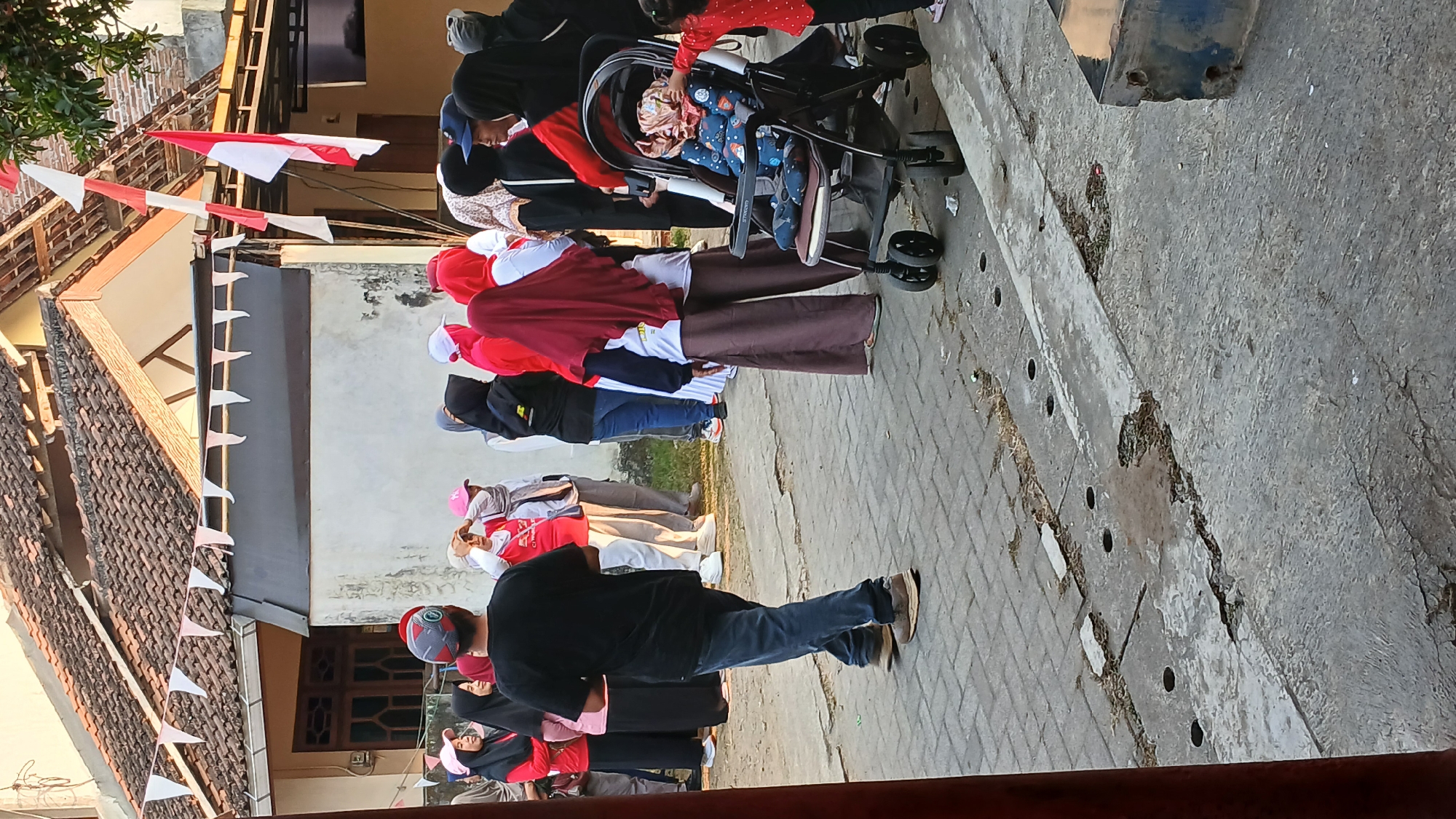 This image depicts a street scene in the city of Mendut, Indonesia, likely during a festive or communal gathering. In the foreground, a person wearing a black t-shirt and blue jeans walks away from the camera. To the right, a stroller with a baby inside is visible, and a small child in a red shirt is reaching towards it. A group of people, mostly women wearing hijabs, are gathered in the middle ground, some wearing red shirts and white or pink hats. Indonesian flags and red and white triangular flags are strung across the scene, suggesting a national celebration. The background shows buildings with tiled roofs and windows. The ground is paved with interlocking bricks and concrete. The overall atmosphere appears to be casual and public.