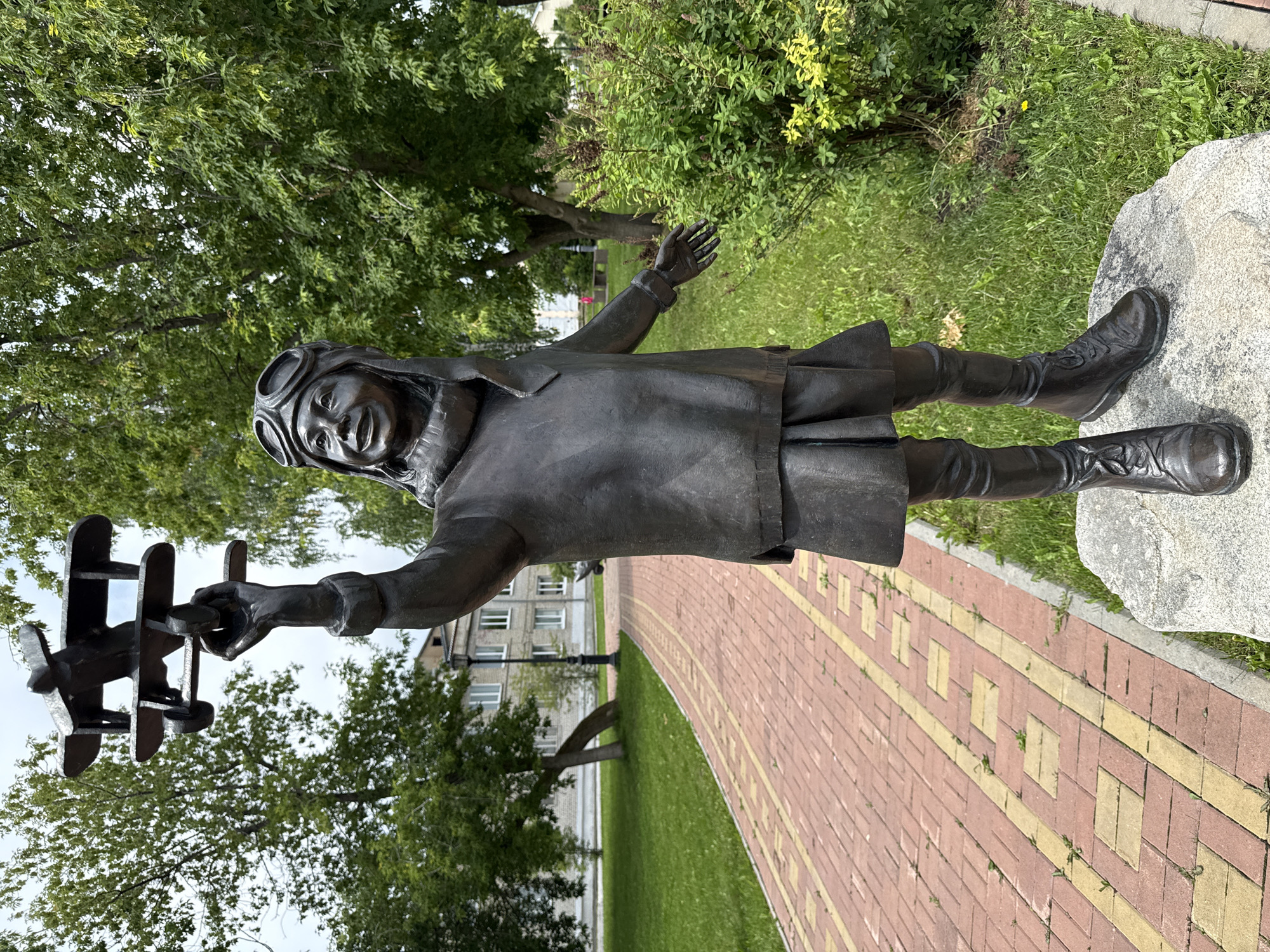 The image depicts a bronze statue of a young girl in aviator goggles and a scarf, holding a model airplane aloft. The statue is situated outdoors on a stone pedestal in a park-like setting. A red brick pathway with yellow accents is visible to the left of the statue, bordered by a manicured lawn. In the background, trees with lush green foliage are present, along with a light-colored building and a lamppost. The statue appears to be located in the city of Krasnoufimsk, Russia, specifically in a public space such as a park or square. The overall scene conveys a sense of youthful aspiration and dreams of flight.