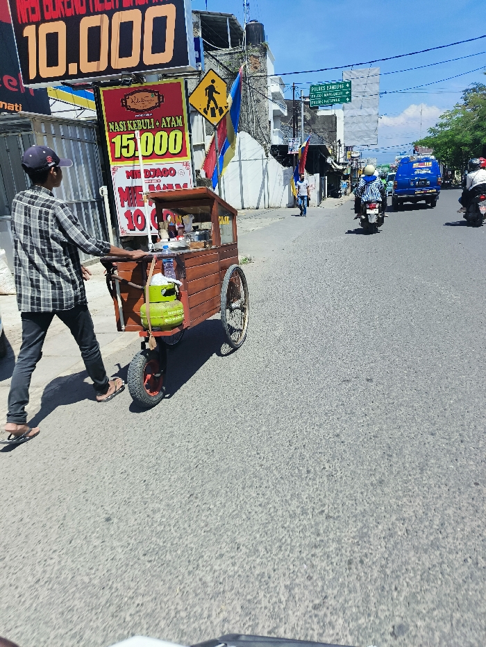 A man wearing a plaid shirt and a black cap is pushing a wooden food cart down a paved road in Margahayu, Indonesia. The cart is loaded with various items, including a green gas tank, a red propane tank, and a metal container. The man is walking on the right side of the cart, with the road stretching out before him. The cart has a sign that says "Nasi Kebuli + Ayam 15,000," and "Mie Django 10,000." There are several other buildings and structures visible in the background, and the sky is clear and blue. The road is relatively empty of traffic, with only a few motorcycles and a blue van driving in the opposite direction. A yellow road sign on the corner warns of a pedestrian crossing. The man is moving to the right and toward the camera. There is another man in the background walking away from the camera on the sidewalk.