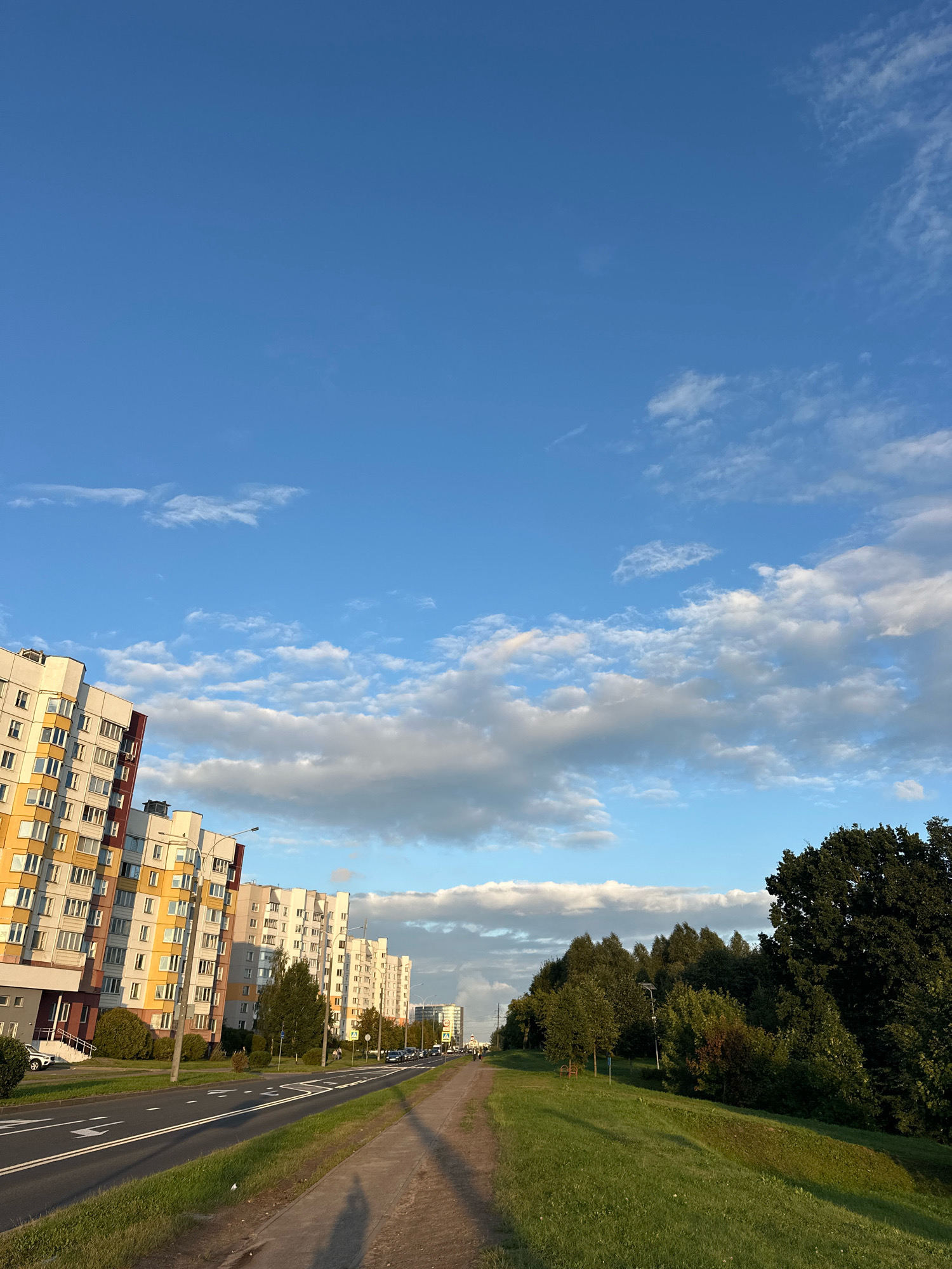 The image depicts a street scene in the city of Minsk, Belarus, likely in a residential area. On the left, a row of modern, multi-story apartment buildings with colorful facades (yellow, white, and pink) lines the street. These buildings have numerous windows and balconies. The street itself is paved with asphalt, featuring double white lines and directional arrows. A sidewalk runs parallel to the street on the right side of the image, adjacent to a grassy embankment. The embankment is covered in green grass and is partially shaded by trees. Beyond the embankment, a dense line of trees forms a green backdrop. The sky is a clear blue with scattered white clouds, suggesting it is daytime, possibly late afternoon due to the lighting and shadows. A few streetlights are visible along the street. In the distance, more buildings and trees can be seen. There are no visible people in the immediate foreground.