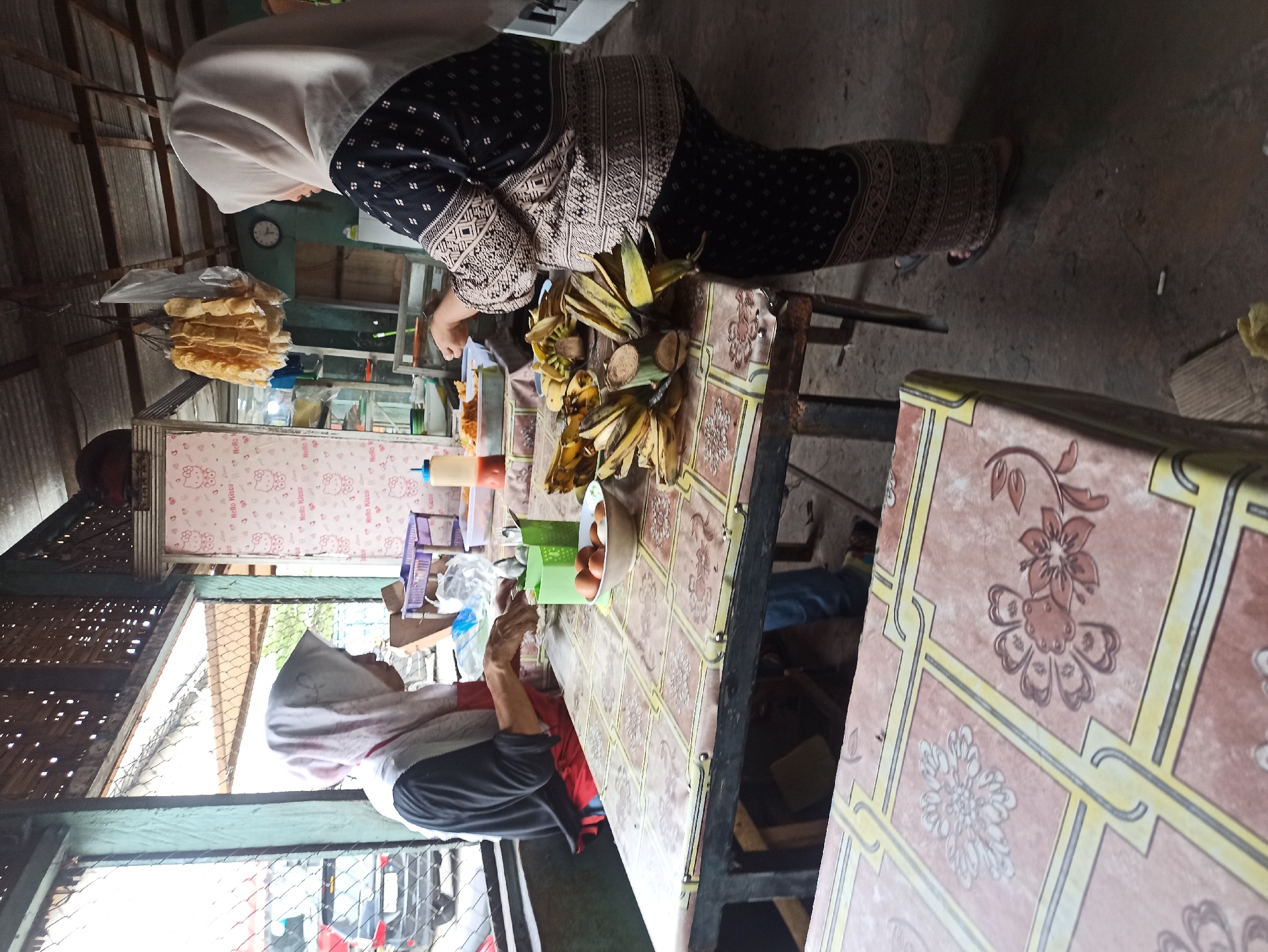 Two women in hijabs are preparing food at a table in an outdoor food stall in Medan, Indonesia. The woman on the left, wearing a red apron over a black shirt and a pink hijab, is slicing something on a plate. The woman on the right, wearing a black and patterned outfit and a beige hijab, is standing and appears to be arranging bananas on a platter. The table is covered with a patterned tablecloth. On the table are also eggs in a bowl, a bottle of sauce, and a purple plastic basket. In the background, there is a car parked outside and a wall decorated with Hello Kitty images. The stall has a tin roof and open sides, suggesting it is a simple, informal eatery.
