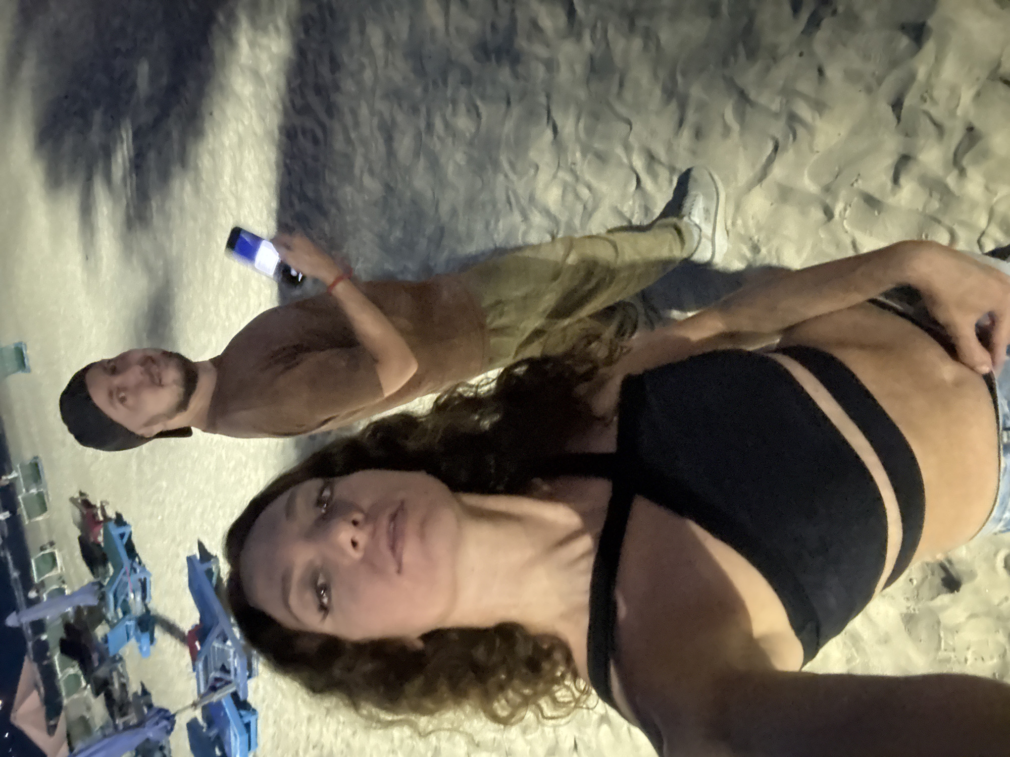A selfie taken on a beach at night. Two individuals, a man and a woman, are visible. The woman is in the foreground, wearing a black top with strappy details and denim shorts. The man is further back, holding a phone. He is wearing a brown t-shirt, olive pants, and a black baseball cap. The background shows sand and beach chairs in the distance, indicating the location is likely a beach in Đà Nẵng, Vietnam.