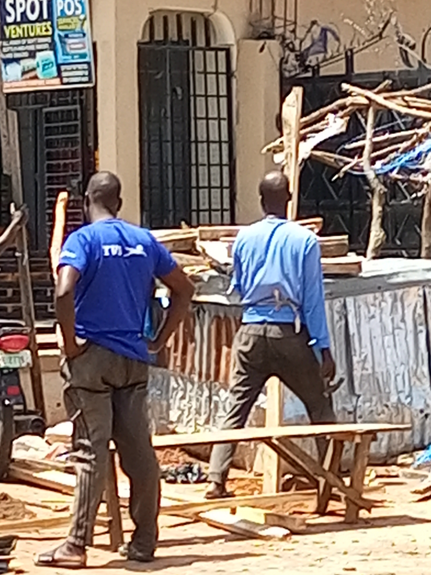 Two men are standing outdoors in front of a building. One man, on the left, wears a blue t-shirt with a logo that appears to say "TVI" with a figure of a swimmer inside a circle. He has his hands in his pockets. The other man, on the right, wears a light blue long-sleeved shirt and dark pants. He has a tool tucked into his waistband. The men appear to be working or observing something in the outdoor area.
The scene suggests an urban or semi-urban environment with elements of construction or disarray. In the background, there is a building with a barred window and an arched entrance. To the right of the building, there is a makeshift structure made of branches and what looks like sheeting or fabric. In the foreground, there is a wooden bench or table with a rustic construction. There are scattered items on the ground, including what appears to be debris and a partial view of a motorcycle on the far left.
A sign is visible on the left side of the building with text that reads "SPOT POS VENTURES" and other unreadable text. The lighting suggests it is daytime, likely under sunny conditions given the shadows. The overall atmosphere appears to be functional and possibly indicative of informal economic activity or a work in progress. There are no clear indications of the time of day or weather conditions beyond the natural light. The location cues are general, showing a building and outdoor space.