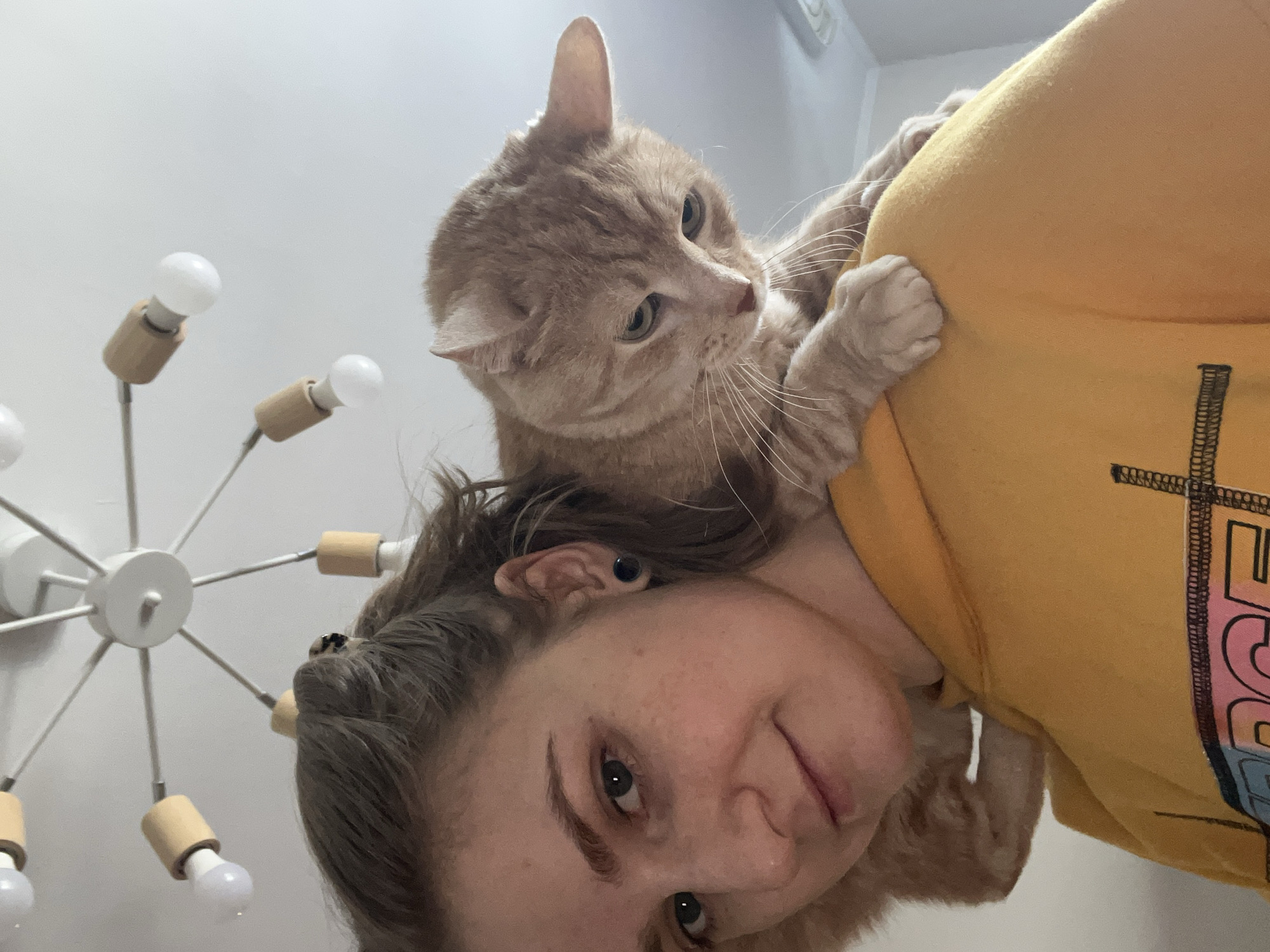 A woman with gray hair is lying down, looking upwards with a slight smile. Perched on her shoulder and head is an orange tabby cat, gripping her yellow hooded sweatshirt with its paws. A modern, white chandelier with multiple bulbs extends from the ceiling in the background. The location cannot be identified as Novosibirsk, Russia from the image.