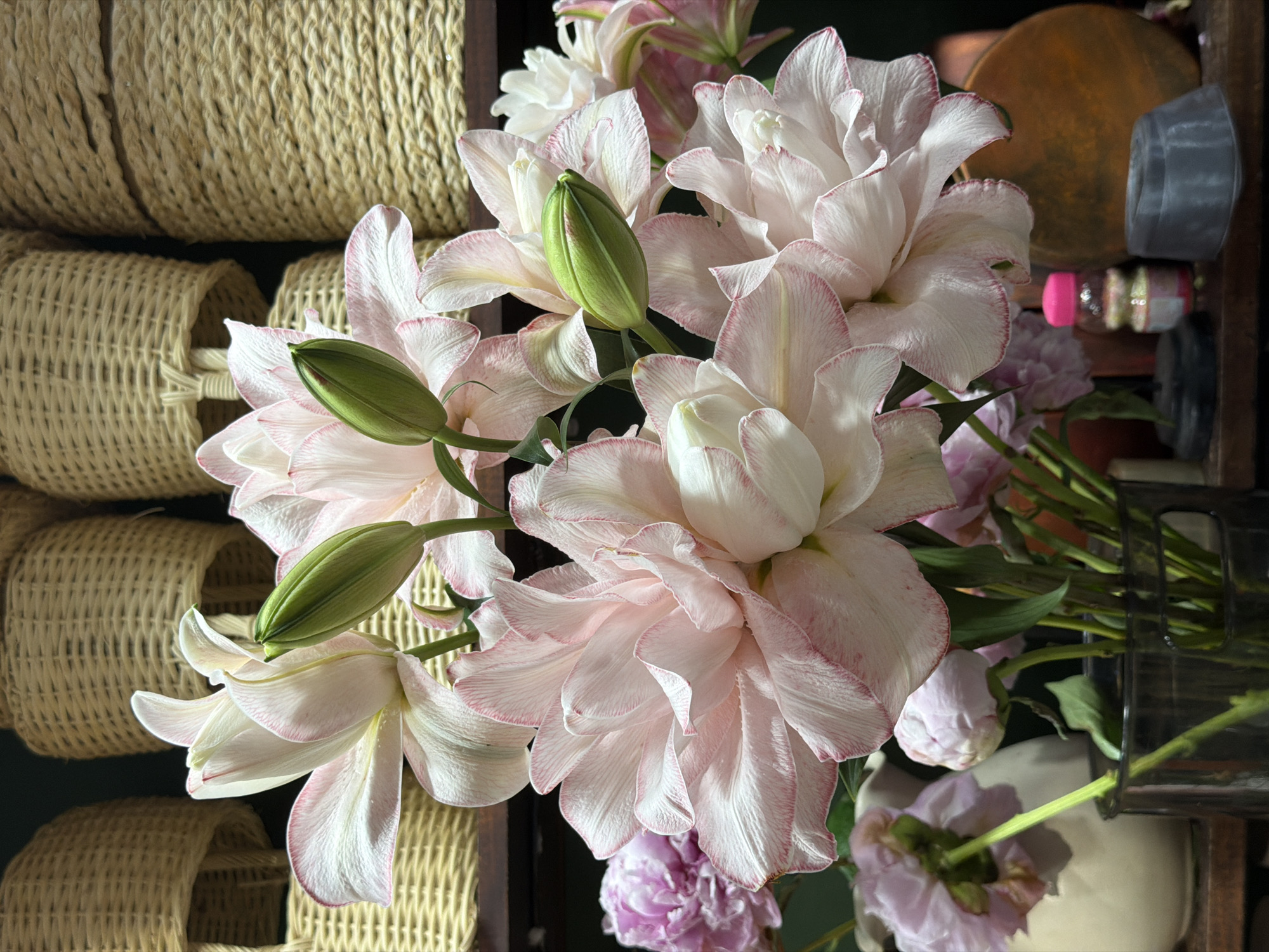 The image shows a close-up of a bouquet of light pink and white lilies with green buds, juxtaposed against a stack of woven baskets. The lilies are the focal point, with their petals exhibiting delicate pink edges. Some purple flowers are visible below. To the right, a blurry background suggests a shelf with various small objects. The baskets are densely woven and appear to be made of a natural material.
The image does not contain information to confirm it was taken in Nha Trang Ward, Vietnam.
