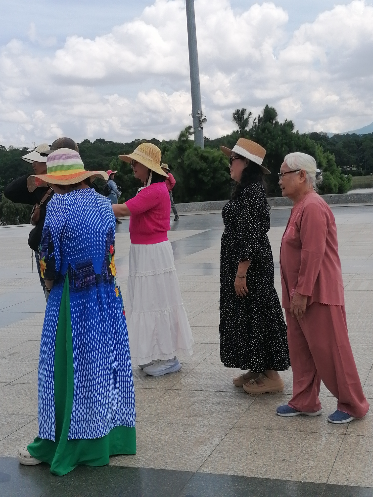 The image depicts a group of five women standing outdoors on a paved area with trees and a body of water in the background, suggesting a park or scenic overlook. The sky is cloudy.
The woman in the foreground on the left is wearing a long, blue, patterned dress with a green underlay, a wide-brimmed hat with colorful stripes, and cream-colored clogs. She is looking away from the camera.
To her right, a woman in a bright pink top and a long white skirt is visible. She is also wearing a straw hat and appears to be speaking to someone off-camera.
Next to her, a woman in a black, polka-dot dress and a tan straw hat is standing, looking to her right.
Further to the right, an elderly woman with white hair, wearing glasses and a pink outfit consisting of a long-sleeved top and loose pants, is standing and looking to her right.
In the far background, a fifth person, possibly a woman, is wearing a denim jacket and can be seen walking away.
The scene appears to be a tourist location, potentially in Da Lat, Vietnam, given the attire of the women (wide-brimmed hats for sun protection) and the presence of lush greenery in the background, which is characteristic of Da Lat's climate and landscape. The paved ground could be part of a plaza or pathway at a landmark like Xuan Huong Lake or a memorial site in the city.