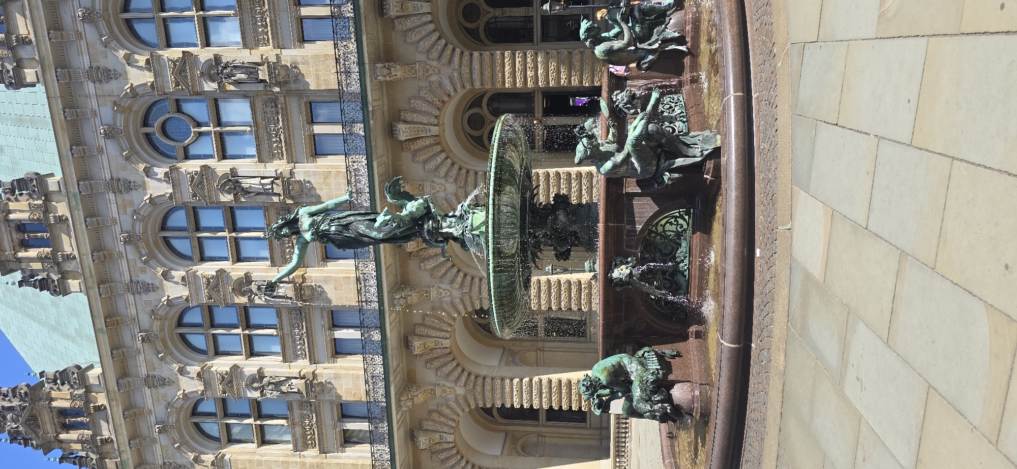 The image shows a bronze fountain in front of a historic building. The fountain features a central sculpture of a woman holding a torch, standing on a winged creature. Water cascades from the central sculpture and overflows into a large basin, which is supported by several smaller sculptures of figures and mythological creatures. The building behind the fountain is ornate, with classical architectural elements, including arched windows, columns, and decorative carvings. The scene appears to be in a public square with paved ground. This fountain is known as the Hygieia Fountain and is located in the Rathausmarkt, the main square in front of the Hamburg City Hall (Rathaus) in Hamburg, Germany. The time of day is sunny, indicating daytime. No people are prominently featured, though a few indistinct figures can be seen in the background near the building.