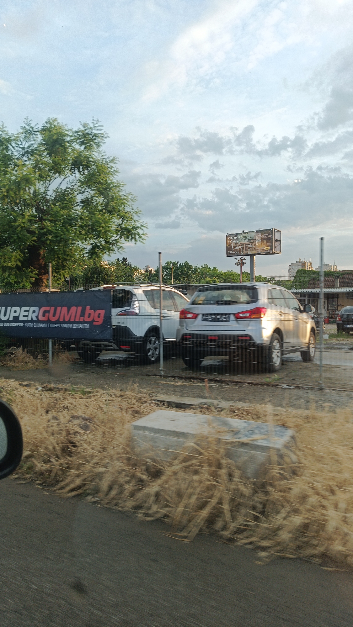 The image captures a roadside view in Burgas, Bulgaria, featuring a fenced-off lot containing several parked cars. A white Ford S-Max and a silver Mitsubishi ASX are visible behind a banner advertising "SUPERGUMI.bg," suggesting the location may be a tire shop or dealership. In the background, a billboard with text and images stands atop a tall pole. Across the foreground, dry, yellow grass and a concrete block are present, blurring with the apparent motion of the camera from a moving vehicle. The sky overhead is cloudy, with a tree partially visible on the left. In the distance, buildings can be discerned. A car's side mirror is visible at the left edge.