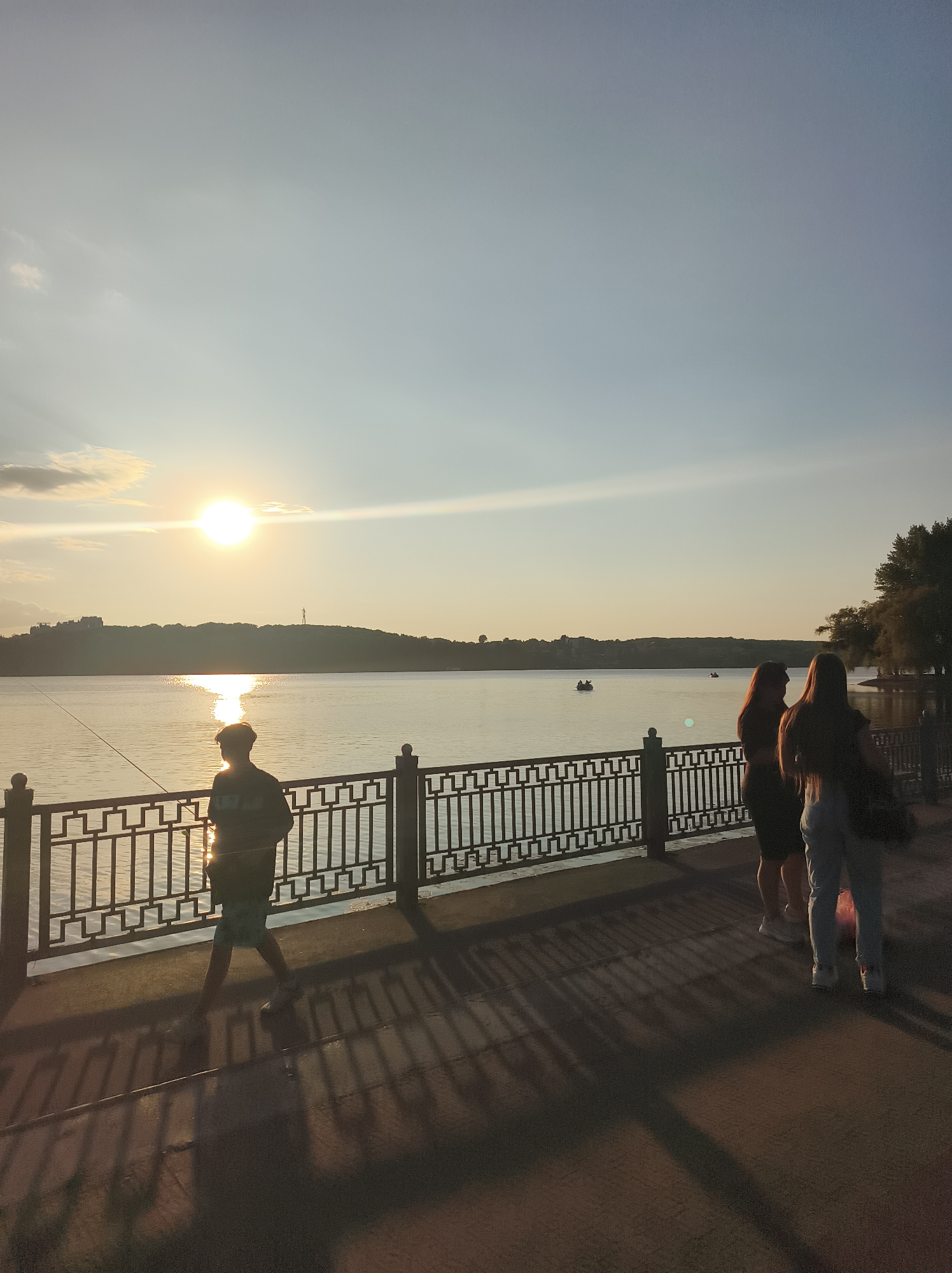 The image depicts a sunset scene by a body of water, likely Ternopil Pond in Ternopil, Ukraine. A lone figure, silhouetted against the setting sun, stands near a decorative metal railing, appearing to be fishing. Further along the railing, three individuals are observed standing together, two women and a child. In the background, a distant tree line forms the horizon of the pond, and several small boats are visible on the water. The ground in the foreground displays the distinct shadows cast by the railing and the people. The sky is a gradient of pale blue and yellow, with the sun prominently positioned near the horizon, its light reflecting on the water.