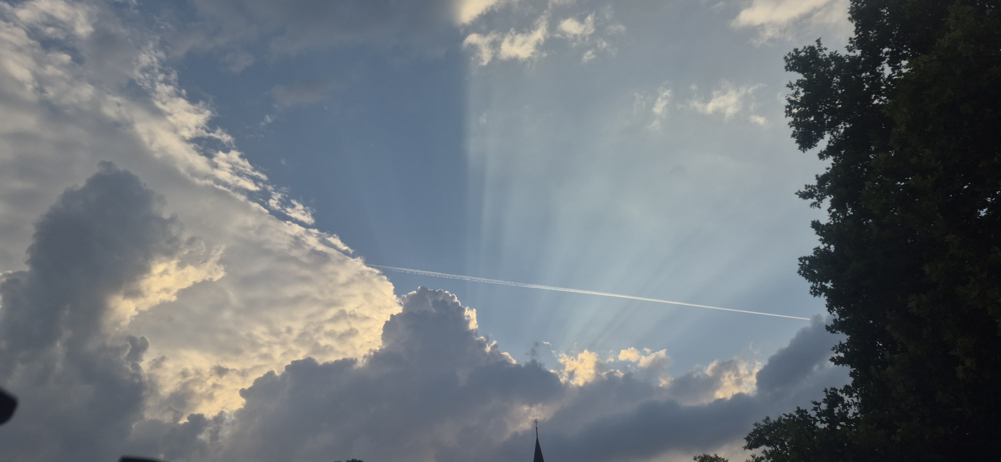 The image captures a dramatic sky over a cityscape, with a large tree on the right side obscuring the view of buildings, except for a church steeple partially visible at the bottom center. The sky is filled with a variety of clouds, some dark and heavy, others illuminated by sunlight, creating visible sunbeams. A contrail from an airplane stretches across the frame. The scene appears to be in Ingolstadt, Germany, based on the presence of a church steeple that is characteristic of European architecture.