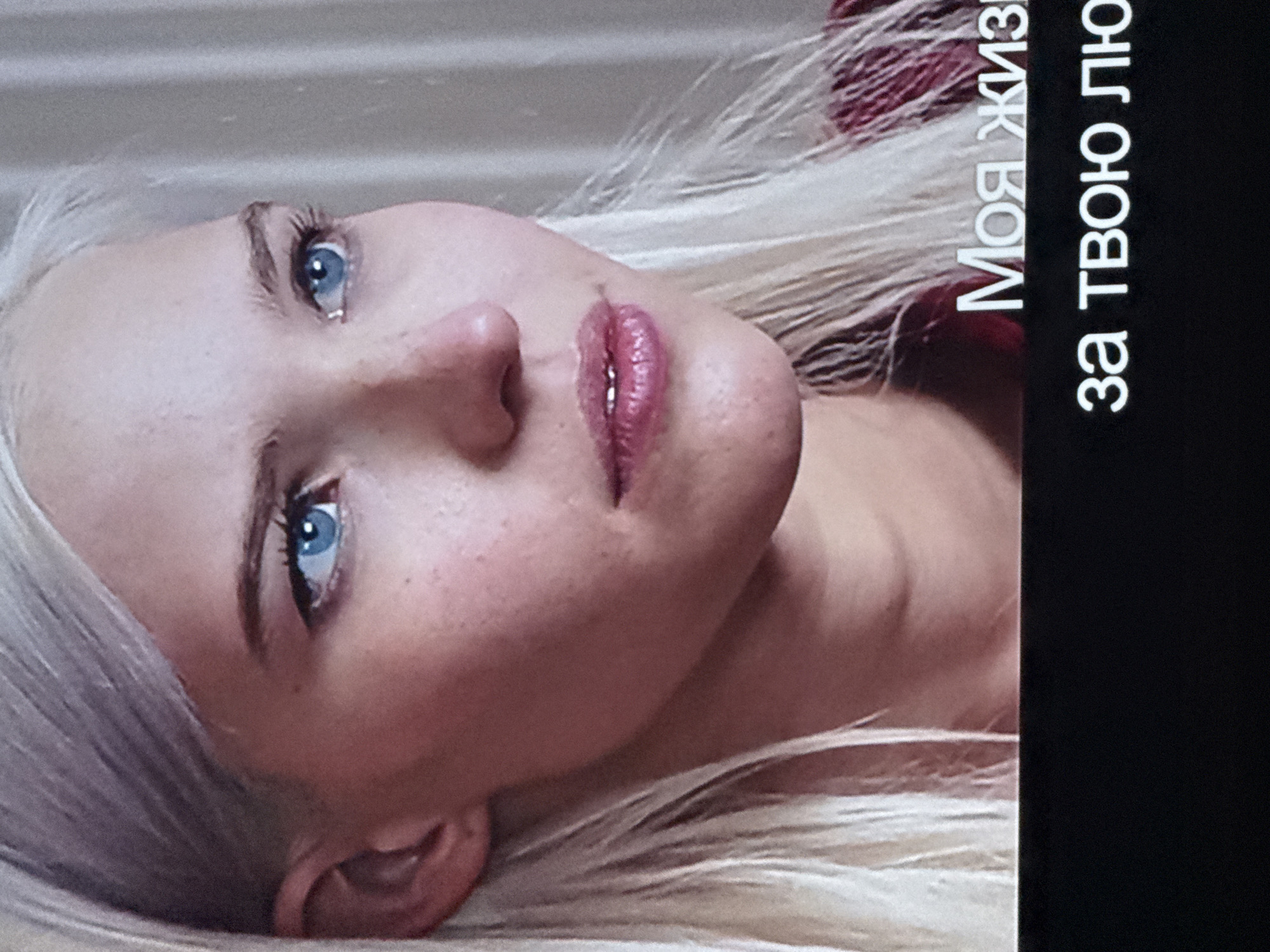 A close-up of a light-skinned woman with blonde hair and blue eyes. She is looking upwards, and her head is tilted to the left. There is some Russian text on the right side of the image. The location is unknown.