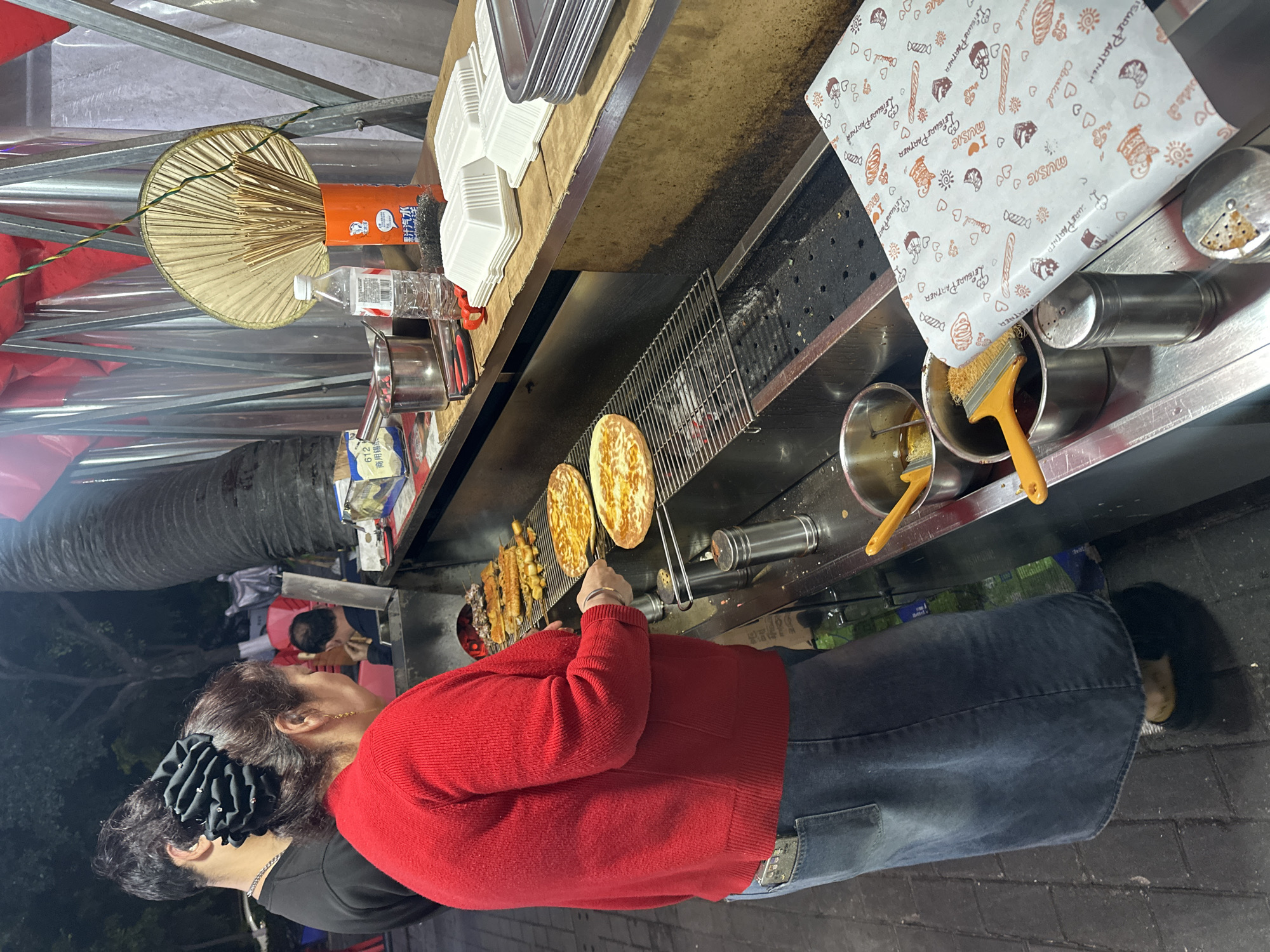 The image depicts a nighttime street food stall in operation, likely within a bustling market environment in the Chongqing High-tech Zone, China.
The main subject is a person, viewed from behind, wearing a bright red long-sleeved sweater and dark blue jeans. Their dark hair is gathered, adorned with a large black ruffled accessory. This individual appears to be a food vendor, actively preparing items on a grill.
The food stall is equipped with a long charcoal grill, visible with glowing embers. On the grill, two round, flatbread-like items (possibly small pizzas or savory pancakes) are cooking, alongside several skewers laden with various meats and vegetables.
Other objects integral to the stall include a traditional round hand-held fan made of bamboo sticks, stored in an orange container. A clear plastic water bottle with "VIMA" printed on its label is also present on the counter. Various cooking utensils, such as brushes resting in metal bowls, and stacks of white disposable food containers, are arranged on the workstation. The counter surface shows signs of regular use.
Notable text includes "LEISURE PARTNER," "MUSIC," "Classical," and "DUZO" printed on the patterned wrapping paper, which features small illustrations of hearts and baked goods. The orange container holding the fan displays "果汁汽水" (Juice soda). A box beneath the counter is labeled with "商用锡纸" (Commercial foil) and the number "612."
The scene conveys the lively ambiance of an outdoor food preparation area during the evening, with the vendor focused on their task. The background is softly blurred, showing glimpses of other market activities and patrons.