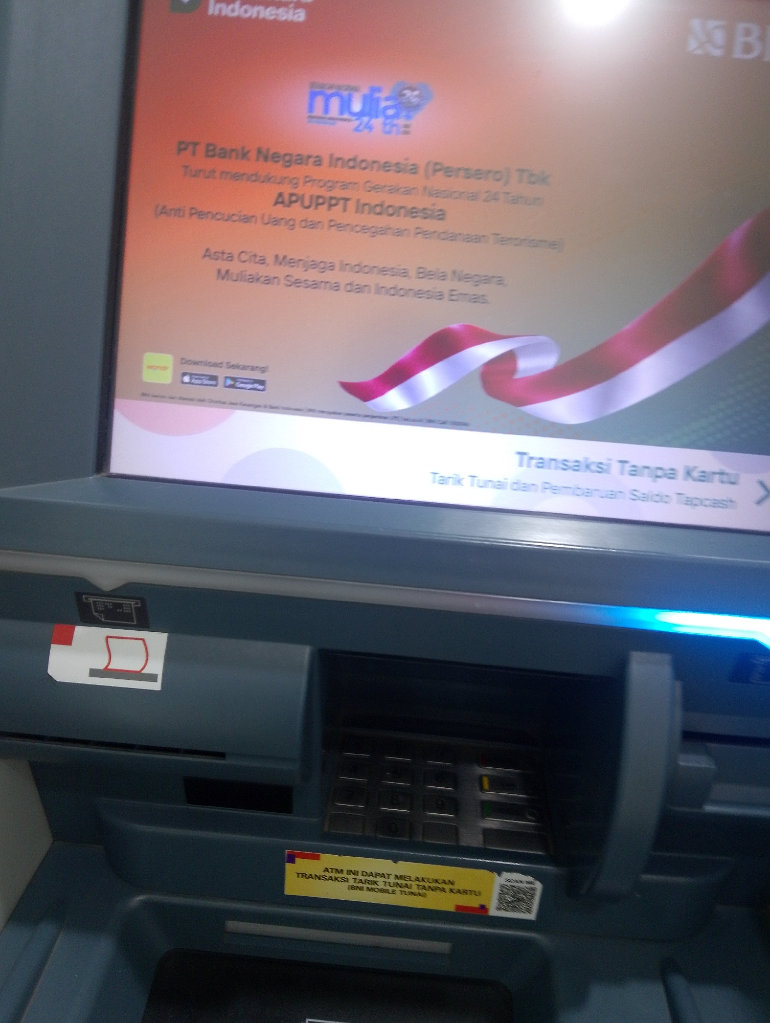 The image shows an ATM screen with a digital display featuring the logo "mullia 24". Text on the screen reads "PT Bank Negara Indonesia (Persero) Tbk Turut mendukung Program Gerakan Nasional 24 Tahun APUPPT Indonesia (Anti Pencucian Uang dan Pencegahan Pendanaan Terorisme) Asta Cita, Menjaga Indonesia, Bela Negara, Mulikan Sesama dan Indonesia Emas." Below this, there is a banner indicating "Transaksi Tanpa Kartu" (Cardless Transaction) with "Tarik Tunai dan Pembaruan Saldo Tapcash" (Cash Withdrawal and Tapcash Balance Update) visible, along with an arrow suggesting further options.
The lower part of the image displays the physical ATM hardware. A card slot is visible with a red and white sticker indicating its purpose. A keypad for PIN entry is present. A yellow sticker with the text "ATM INI DAPAT MELAKUKAN TRANSAKSI TARIK TUNAI TANPA KARTU (BNI MOBILE TUNAI)" (This ATM can perform cardless cash withdrawal transactions (BNI MOBILE CASH)) and a QR code for scanning is affixed below the keypad. The overall environment appears to be an indoor public space, likely a bank or a commercial area in Serbelawan, Indonesia, given the language and banking context. No individuals are present in the image.