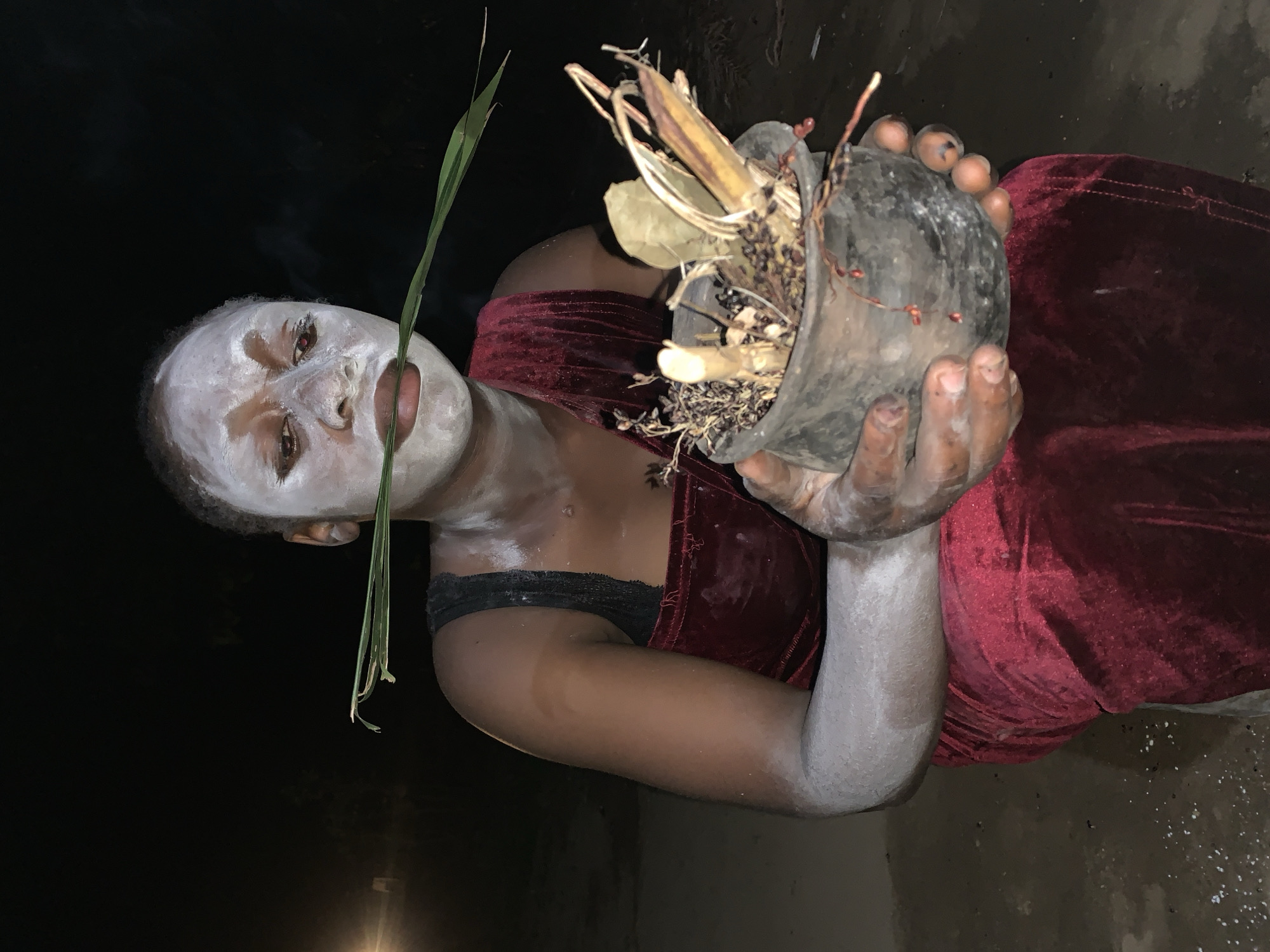 A medium shot depicts an individual with dark skin, presumably female, facing the viewer with their face painted white. A single blade of green grass is held in their mouth. They are wearing a dark red, velvet-like top with black lace trim visible at the neckline. The person is holding a dark-colored pot filled with dried leaves, twigs, and other organic matter. Their arms are also covered in white powder.
The setting appears to be outdoors at night. In the background, a dim light source is visible. The location is cited as Agban, Nigeria, though there are no specific landmarks visible in the image. The ground beneath appears to be wet or sandy.