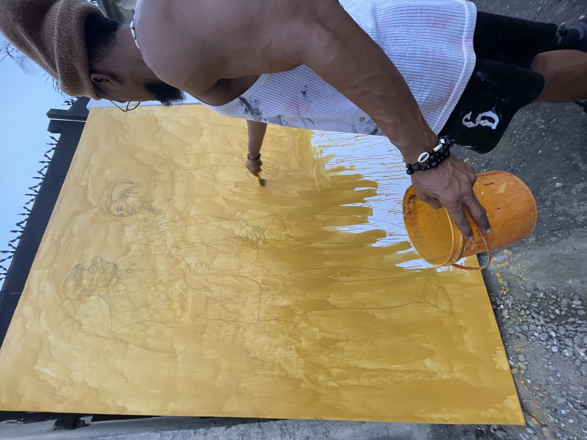A man wearing a brown beanie, glasses, a white tank top, and black shorts is painting a large, rectangular wooden canvas with an outline of two people on it. He is pouring a yellow liquid from an orange bucket onto the canvas and also using a paintbrush. The scene appears to be outdoors in Ado, Nigeria, as the painting is taking place on a concrete surface with small rocks and debris scattered around. The canvas is leaning against a black metal fence. The man is wearing a beaded bracelet on his left wrist and a necklace around his neck.