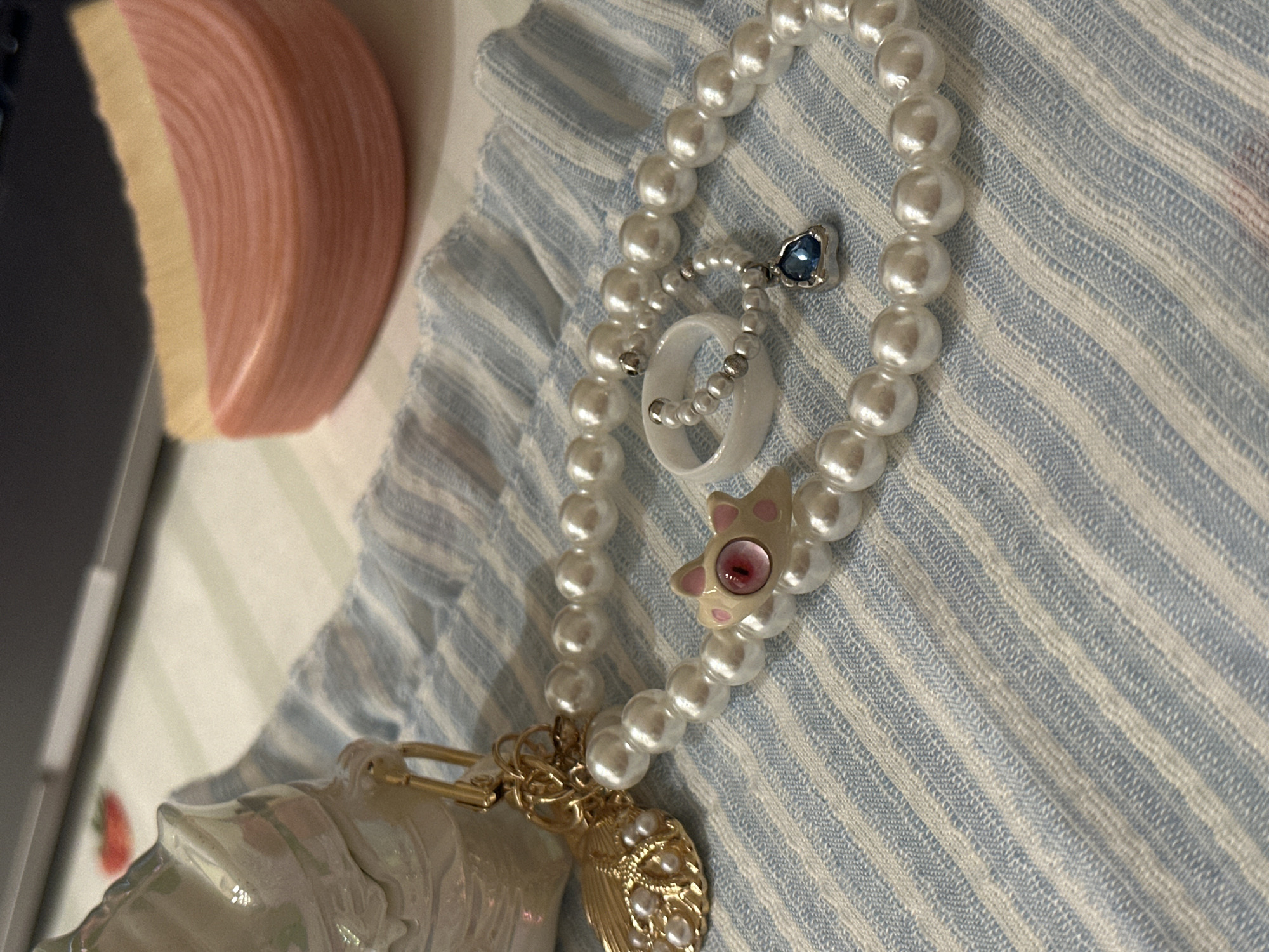 The scene, located in Bar, Montenegro, displays a static arrangement of various items positioned on a light blue and white striped fabric surface. In the midground, a strand of approximately 6-8mm white, spherical pearls is laid out in a curved formation. A cream-colored, stylized cat-head charm, featuring pink detailing on its ears and a central red eye with a dark pupil, is situated on this pearl strand. Adjacent to this, a smooth, plain white ring rests on the fabric. A smaller, finer bracelet, composed of approximately 3-4mm white pearls interspersed with silver-colored beads, is looped around the white ring. This smaller bracelet suspends a silver-toned pendant encasing an irregularly shaped, faceted blue gemstone. To the left, a pearlescent white decorative object, possibly a stylized shell or horned creature, is connected via gold-colored metal hardware to a gold-colored charm, resembling a plume or feather, which is embedded with multiple small white pearls. The fabric exhibits a woven texture with visible striations and a ruffled edge is present on the right side. In the blurred background, a dark gray rectangular object, consistent with an electronic device, is partially visible at the top. A small red object, resembling a strawberry graphic, is visible on a light surface behind the pearlescent item. In the upper right background, a semi-circular, layered object displaying pink and pale yellow coloration, consistent with a bar of layered soap, is present.