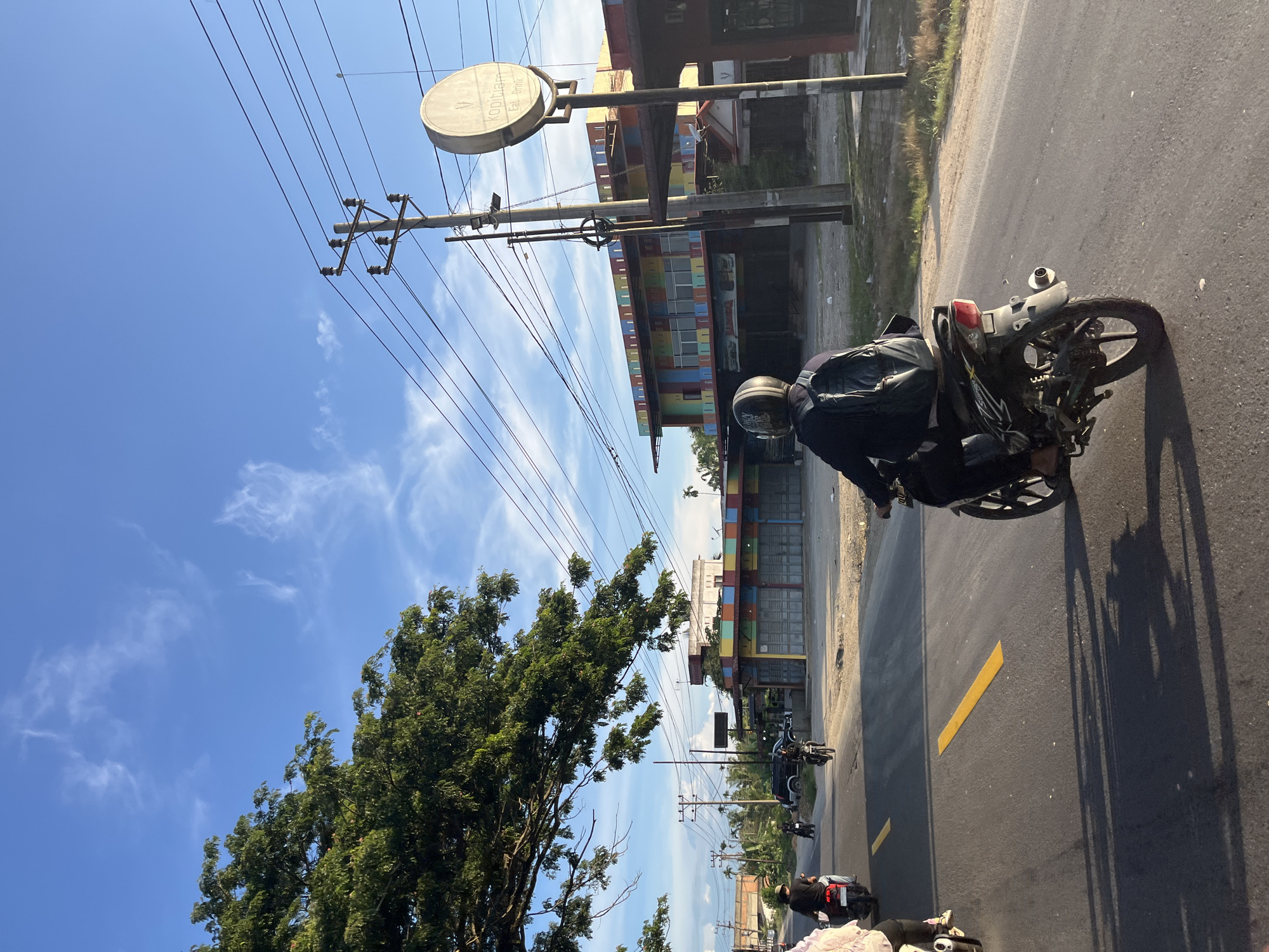 This image captures a street scene in Seuneubokpunti, Indonesia, under a clear blue sky with scattered wispy clouds. The sun is casting long shadows, indicating it is likely late afternoon. In the foreground, a person is riding a motorcycle, seen from behind, wearing a dark jacket and a helmet, with a black backpack. The motorcycle is black and has visible markings. The road is asphalt with yellow lane markings. Several other motorcycles are visible further down the road, also with riders.
To the right of the road, a building with a colorful facade, featuring red, blue, yellow, and green panels, is present. Above the building, a circular sign with text that appears to read "kopitiam" and "Eat Drink" is mounted on a pole. Utility poles with numerous wires crisscross the sky. On the left side of the image, a large tree with dense green foliage extends over the road. In the background, more buildings and palm trees can be seen. The overall impression is a typical street in a town or city in Indonesia.