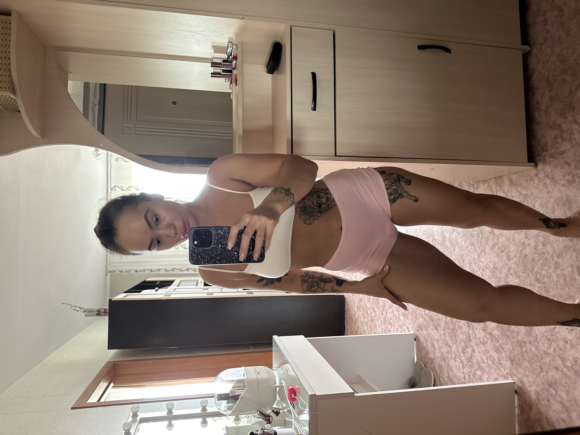The image shows a woman taking a mirror selfie in what appears to be a bedroom or dressing area. She is wearing a white sports bra and pink shorts. She has tattoos on her arms, legs, and torso. The room has a vanity with makeup brushes and a makeup organizer to the left, and a wardrobe with shelves and drawers to the right. There is a mirror on the wardrobe. The flooring is a pink speckled pattern. The background includes a door and a window with curtains. The image was taken in the city of Yuzhnouralsk, Russia.