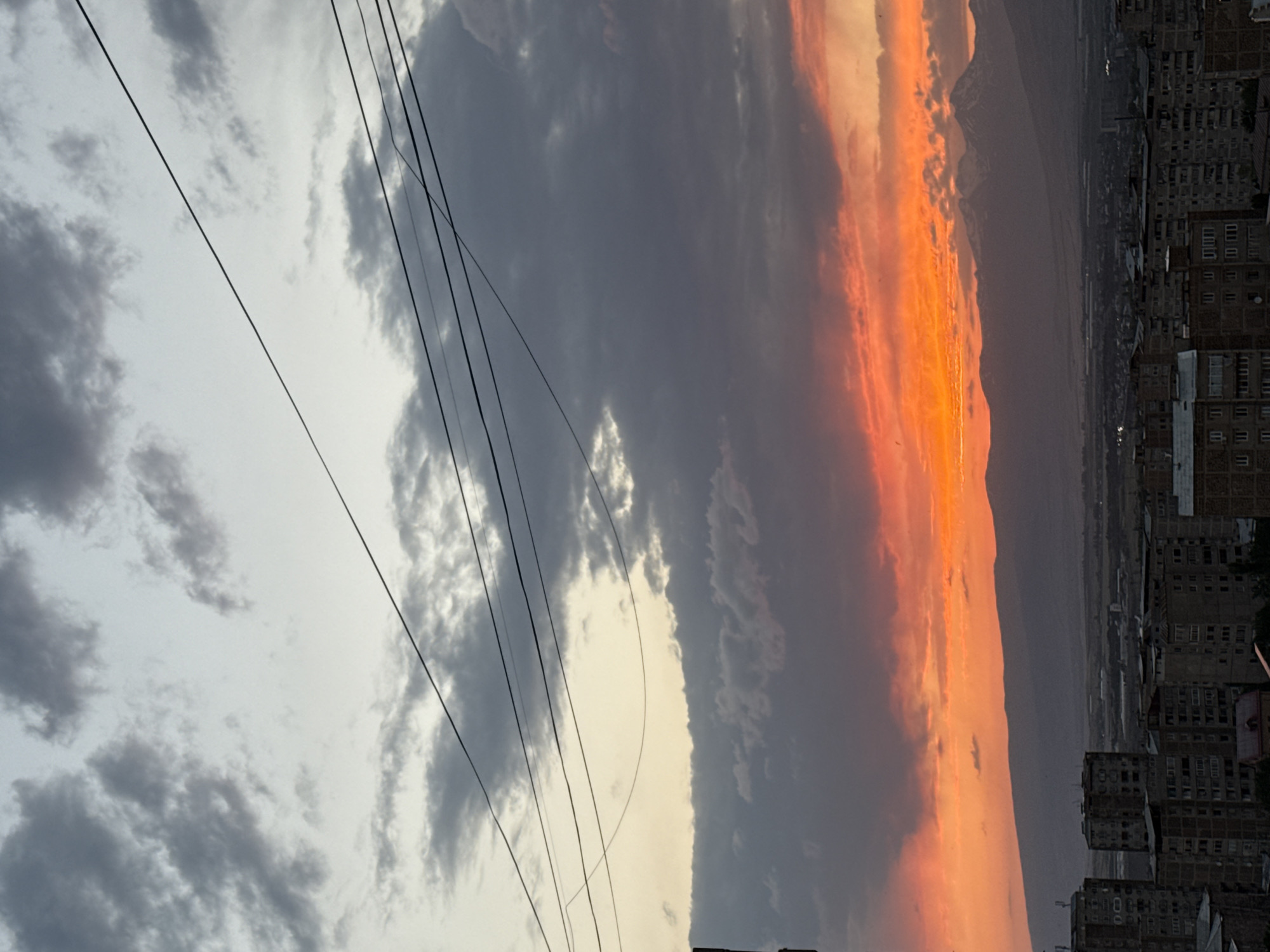 This image depicts a panoramic view of the city of Abovyan, Armenia, during what appears to be sunset. In the foreground, dense residential buildings with a Soviet-era architectural style dominate the urban landscape. These structures are characterized by their multi-story designs and uniform facades.
Beyond the city, a range of mountains forms a dramatic backdrop. The peaks of these mountains are dusted with snow, suggesting higher altitudes and possibly cooler temperatures. The sky above is a canvas of dramatic cloud formations, transitioning from shades of gray and white at the top of the frame to a vibrant orange and red gradient near the horizon, indicative of the setting sun. Several utility wires are visible crisscrossing the sky, adding a man-made element to the natural grandeur. No people or animals are discernible in the image. The overall scene conveys a sense of urban life set against a majestic natural environment, illuminated by the striking colors of twilight.
