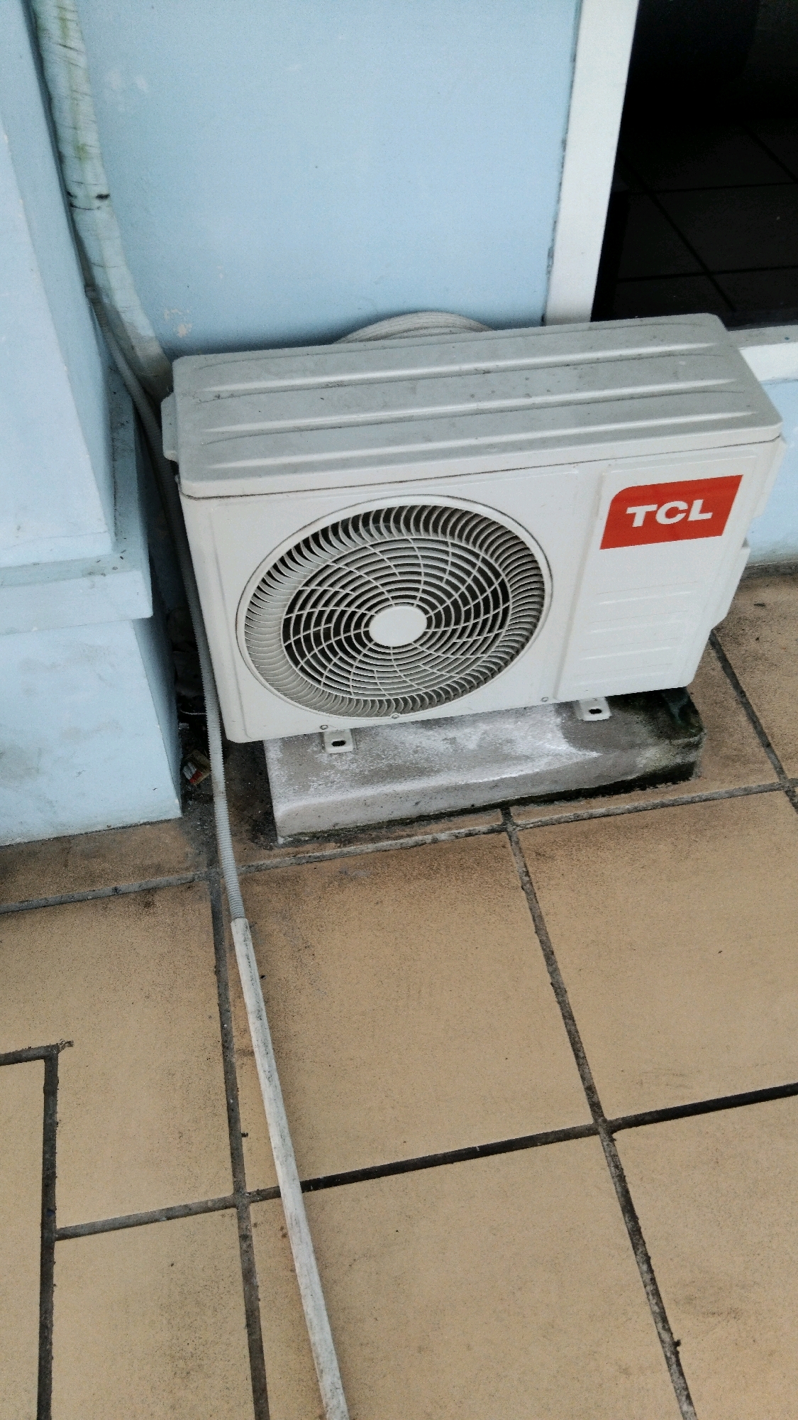 An outdoor air conditioning unit, with the TCL logo visible, sits on a concrete base on a tiled floor. The unit is white and has a circular fan grille on its front. A white conduit runs vertically from the unit down towards the ground. The wall behind the unit is painted light blue. The scene is likely in Pekalongan, Indonesia, based on the provided information.