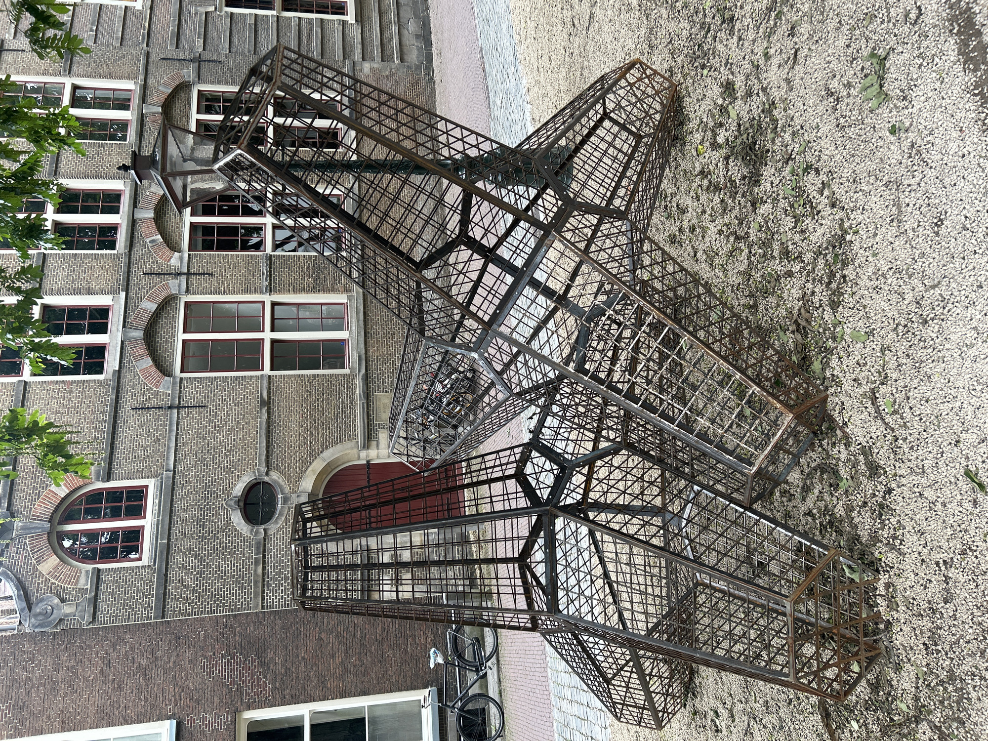 The image shows a metal sculpture composed of geometric cages in front of a brick building. The sculpture is made of rusted metal bars and is in a state of disrepair or is intentionally rustic. It is positioned on a ground covered with small white pebbles and some scattered leaves. To the left of the sculpture, a bicycle with a basket is parked on a cobblestone path. Behind the sculpture, the building features arched windows with red frames, and a red door. Trees with green leaves are visible extending into the frame from the top. The architecture of the building suggests it is an older structure, possibly historical. The overall scene appears to be outdoors, in a public space. This photograph was taken in Leiden, Netherlands, specifically in the courtyard of Museum De Lakenhal.