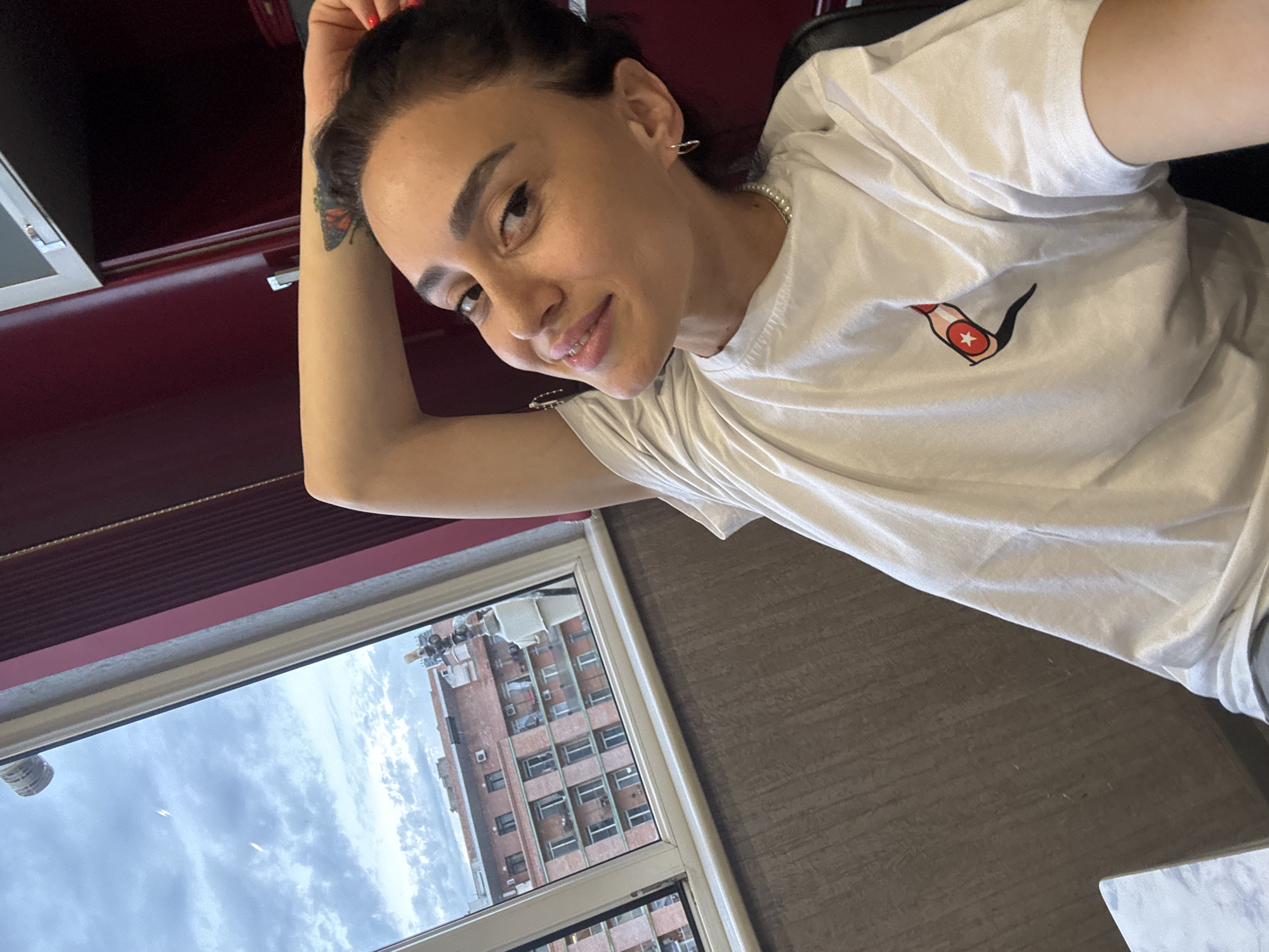 A woman is taking a selfie indoors, with her right arm behind her head. She is wearing a white t-shirt with a red and white emblem on it, a pearl necklace, and hoop earrings. Her hair is dark and pulled back. She has a butterfly tattoo on her left forearm. The background shows a window with a view of apartment buildings and a cloudy sky. The room appears to be an office or bedroom with dark red cabinetry and a textured brown wall. The window frame is white and the glass reflects some of the interior. The overall lighting is natural and diffused.