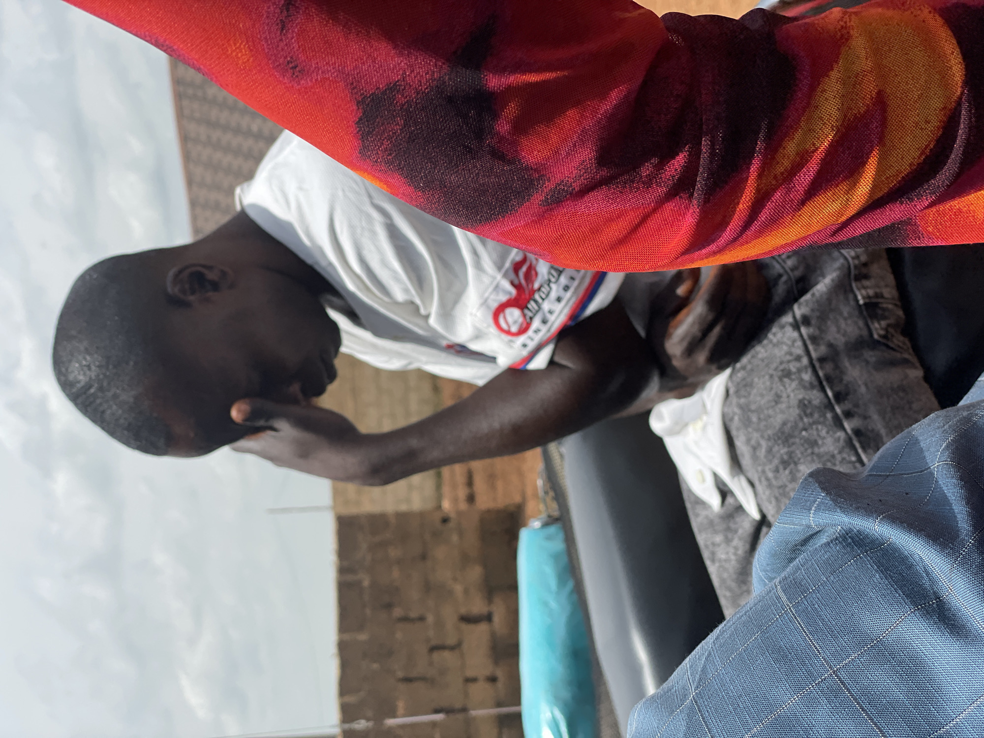 In the city of Kassa, Nigeria, a man with dark skin and short hair sits with his head bowed, his right hand pressed against his forehead. He is wearing a white t-shirt with a circular logo on the left side and a white cloth draped over his lap. To his right, a portion of a person's arm is visible, adorned with a red and orange tie-dye patterned fabric. The background consists of a brick wall and a partially visible white and blue object. The man appears to be in a contemplative or distressed state, possibly outdoors given the bright sky and the general outdoor ambiance suggested by the brick wall and open sky. The overall impression is a candid moment captured, focusing on the man's posture and the textures of the clothing.
