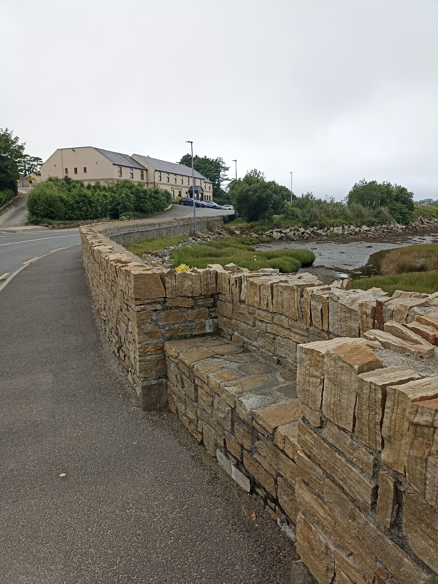 This image captures a scenic view of Annagry West, Ireland, under an overcast sky. The foreground is dominated by a rustic stone wall, meticulously constructed with rough-hewn, light-brown stones. The wall follows the curve of an asphalt road, which leads away from the viewer and into the distance. To the left of the road, a grassy embankment is visible.
Beyond the road and the wall, the scene opens up to a tidal estuary with mudflats, bordered by lush green vegetation and reeds. In the mid-ground, a long, beige building with a dark roof stands prominently, identified by visible text as a "HOTEL." Several parked cars are visible in front of the hotel, suggesting it is open for business. A streetlamp stands near the hotel entrance.
The overall atmosphere is one of tranquility and natural beauty, enhanced by the muted light of the cloudy day. The presence of the hotel hints at tourism in the area. The natural landscape of the estuary and the traditional stone wall evoke a sense of place specific to coastal Ireland. There are no people visible in the image. The time of day appears to be daytime, given the ambient light.