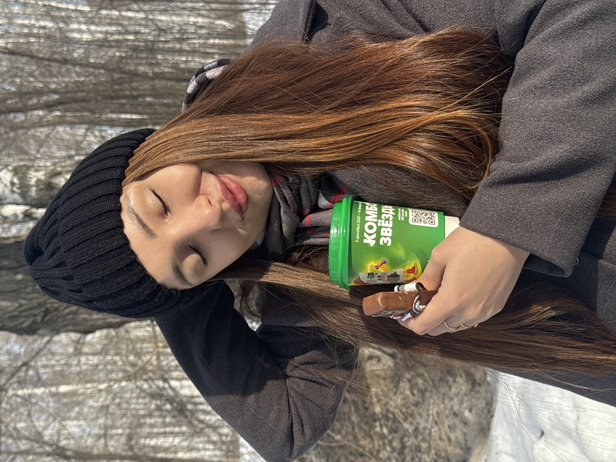 An individual, appearing female with long reddish-brown hair, is depicted outdoors in a snowy, wooded environment in the city of Chaikovsky, Russia. She wears a black ribbed knit beanie, a dark charcoal-grey winter coat, and a grey and red plaid scarf. Her eyes are closed, head tilted slightly, and lips display a subtle smile. Her right hand is positioned near the back of her head. In her left hand, she holds a green disposable cup with a green lid, which displays white text including "2 декабря 2023 - 15 февраля..." and "КОМБО ЗВЁЗДЫ," alongside a QR code. Also held in her left hand, partially unwrapped, is a brown chocolate bar. The background shows bare trees, consistent with a winter or early spring setting, with snow covering the ground under bright sunlight.