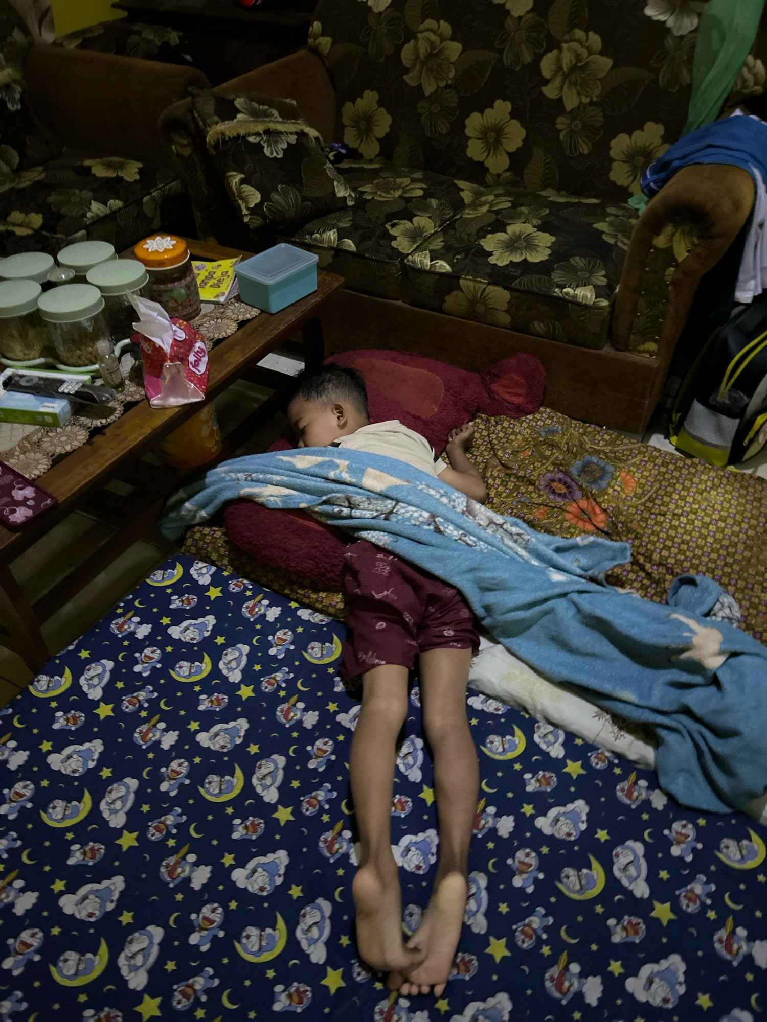 In a dimly lit room in Cihaurbeuti, Indonesia, a young boy lies asleep on the floor. He is covered by a blue blanket and a darker-colored throw. The floor is covered by a blue sheet with a repeating pattern of cartoon characters, moons, and stars. To the left of the boy, a wooden table holds several jars, a packet of tissues, a small yellow book, and a blue container. Behind the boy, a large reddish-brown teddy bear is visible, and further back, a floral-patterned sofa occupies the background. The overall impression is of a domestic, lived-in space.