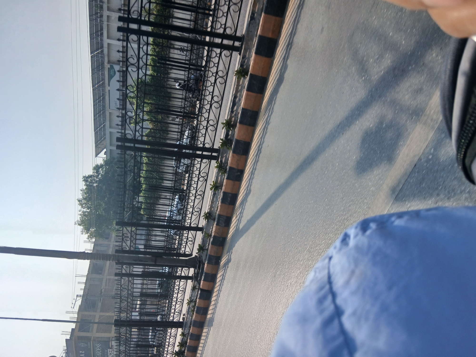The image depicts a street scene in Peshawar City Tehsil, Pakistan, likely taken from a moving vehicle. In the foreground, a portion of a blue garment is visible, suggesting the vantage point is from within a car or bus. The street is paved and shows some shadow patterns, indicating sunlight.
Behind the street, a black ornamental metal fence runs horizontally across the image. Beyond the fence, a row of small, green plants is visible. Further back, there is a paved area with a yellow and black striped curb. Several vehicles, including cars and motorcycles with riders, are seen on the other side of the fence.
In the background, a multi-story building is under construction, with exposed concrete and rebar. Above this, trees are visible. To the right, another building with a large array of solar panels on its roof is present. The sky is clear and bright, suggesting a daytime setting. Text on the buildings is partially visible, with "GARDENS" and "NCER" being discernable on the construction site building. The solar panel building has visible branding, including "ptcl". The overall impression is that of a busy urban environment.