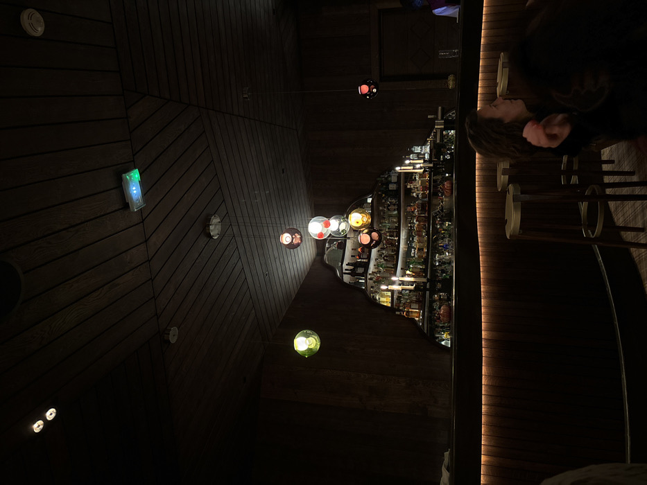 This media file captures the interior of a dimly lit, stylish establishment, likely a bar or lounge, with a warm and intimate atmosphere. The image is rotated 90 degrees clockwise.
The scene features a modern design dominated by dark wooden paneling on the ceiling and walls, arranged in a striking geometric pattern of angled planks. Scattered across this wooden surface are various light fixtures: several small, recessed spotlights, a circular smoke detector-like device, and a rectangular emergency light or control panel emitting a green glow.
A central element of the visual composition is a cluster of modern pendant lights hanging at varying heights. These lights feature spherical glass shades in a diverse palette of colors including red, amber, clear, green, and a muted brown or grey, casting soft, colorful glows.
On the right side of the image (which would be the floor and wall if the image were correctly oriented), a well-stocked bar is visible. Numerous bottles of spirits and liqueurs are neatly arranged on shelves, illuminated from within or by overhead lights, creating bright reflections. The bar counter itself features a contemporary design with a warm, illuminated strip running along its length. Several elegant bar stools with dark seats and light-colored bases are positioned in front of the counter.
One person is visible on the far right, seated at or standing near the bar. Only their head and upper torso are visible in profile, showing dark, possibly curly hair. They appear to be looking forward or slightly downward, suggesting a moment of quiet contemplation or waiting.
The overall impression is one of a sophisticated and cozy evening setting, characteristic of a high-end lounge or restaurant. The dim, colorful lighting and dark wood create an inviting ambiance. No specific activity beyond being present at the bar is distinctly captured, and no discernible text is visible within the frame. The time of day is clearly evening or night due to the reliance on artificial lighting. The location context indicates Val-d'Isère, France.