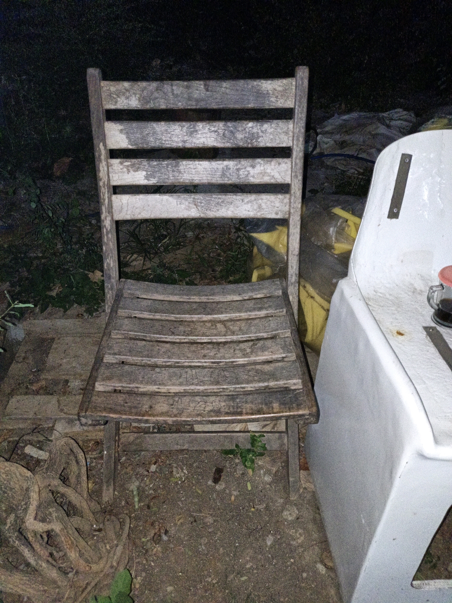 A weathered wooden chair is positioned outdoors at night, next to a white, basin-like object. The chair has a slatted backrest and seat, and its legs are visible, planted on the uneven ground. The ground is a mixture of dirt, small stones, and some greenery, with what appears to be exposed tree roots to the left of the chair. In the background, there are dark shadows, foliage, and some yellow and gray plastic sheeting. The white object next to the chair is smooth and curved, with metal fixtures visible on its surface and a small glass with a red lid containing a dark liquid placed on its edge. The scene appears to be in a somewhat unkempt outdoor area, possibly a backyard or storage space. The location in Cikal, Indonesia is not visually evident from the image.