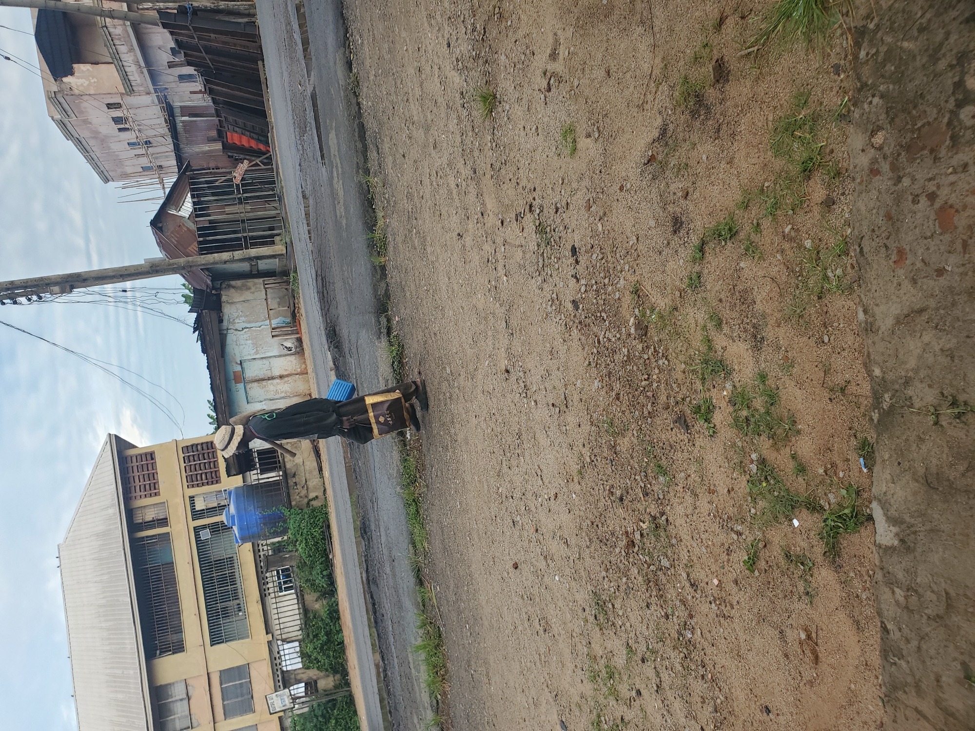 A person is walking on a partially paved road with a dirt shoulder. The road is situated between buildings of varied construction and age in Unknown, Unknown. The person carries a blue container on their head and a box with brown and gold accents. The buildings are multi-story and show signs of weathering and repair. The sky is overcast.