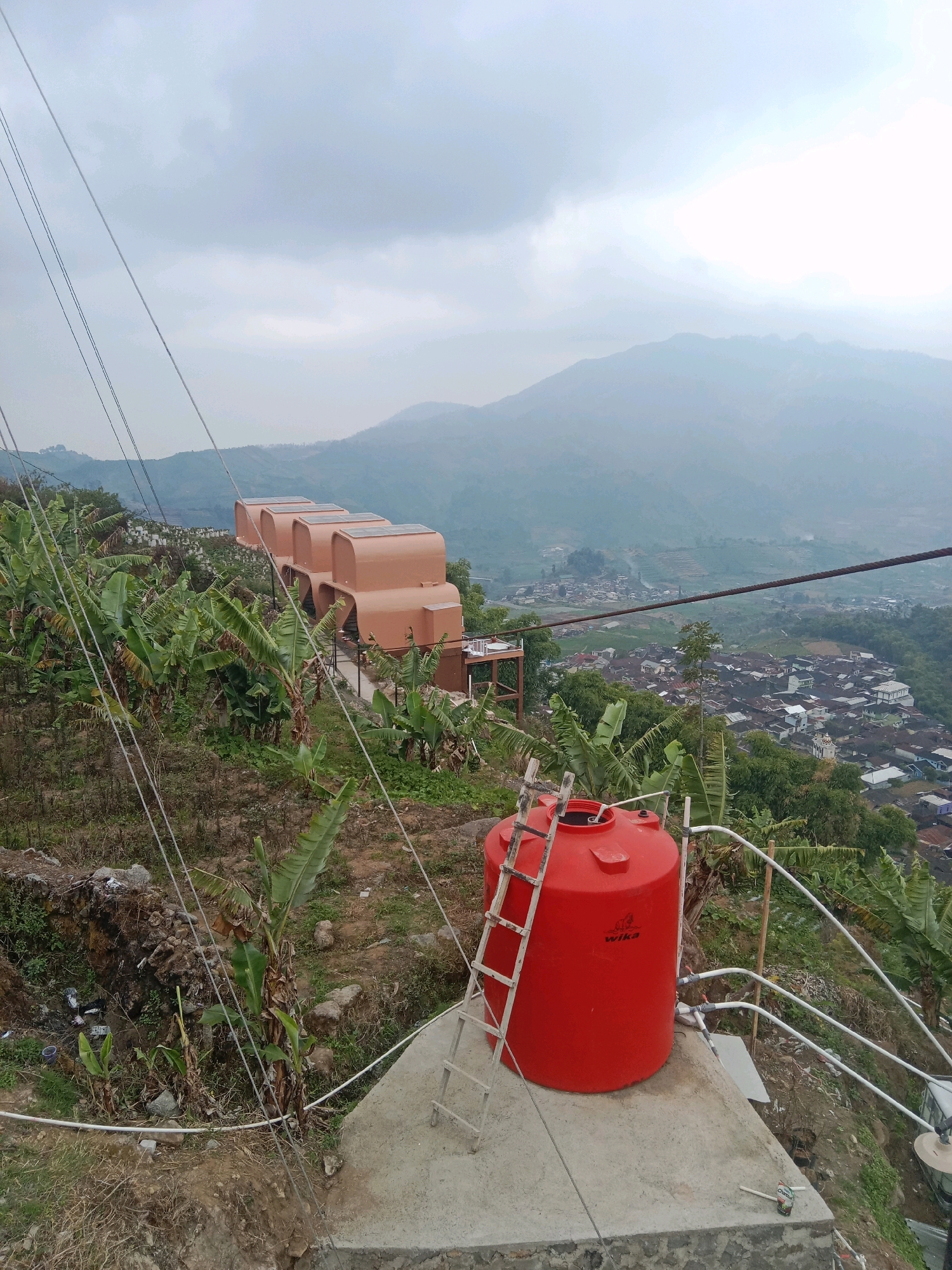 An outdoor shot captures a red water tank with a white ladder leaning against it, situated on a concrete platform. The tank bears the "wika" logo. In the background, several identical, light brown, pod-shaped structures are lined up on a hillside, adorned with what appear to be solar panels on their roofs. These structures are surrounded by lush green banana trees. Beyond them, a hazy vista unfolds, revealing a sprawling village nestled within a valley, with mountains fading into the distance under a cloudy sky. The scene is bisected by a network of overhead wires, contributing to the overall ambiance of a remote, possibly rural, location in Tieng, Indonesia.
