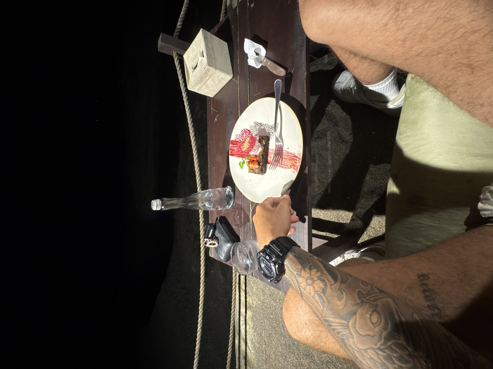 The image presents an overhead view of a partially illuminated outdoor setting. A dark background suggests nighttime or a dimly lit area. A person wearing a black wristwatch on their left arm, which features prominent tattoos, is holding a fork and appears to be eating a dessert on a white plate with a red decorative element. The dessert appears to be a slice of cake with a strawberry. A knife rests near the plate. A tissue box sits near the plate, and there are two empty glass bottles on the side of the table. Another person wearing white sneakers, socks, and light-colored shorts sits with their legs and thighs within the frame. The setting appears to be a wooden table that is a part of a structure supported by ropes and beams, which could be part of a restaurant, bar, or some other outdoor venue, that is consistent with the city of Munggu, Indonesia.