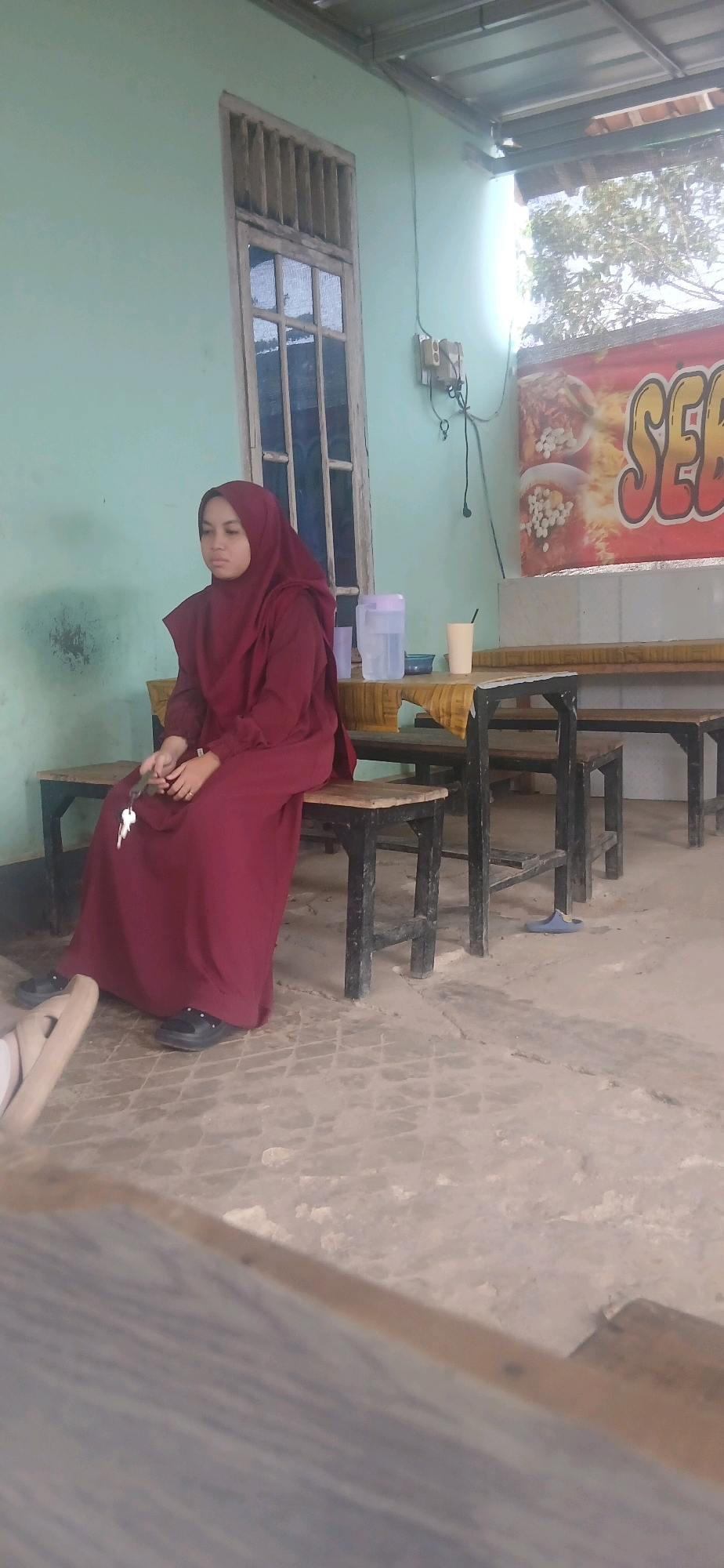 A young woman wearing a maroon hijab and dress sits on a wooden bench in an outdoor covered area. She is holding a set of keys in her right hand. The bench is part of a table and chair set. In the background, there is a light blue wall with a window and an electrical outlet. A banner advertising food is visible on the right side of the image. Several plastic jugs and cups are on a table behind the bench. The ground is concrete. This scene is likely located in Metro City, Indonesia, possibly at a food stall or casual eatery.