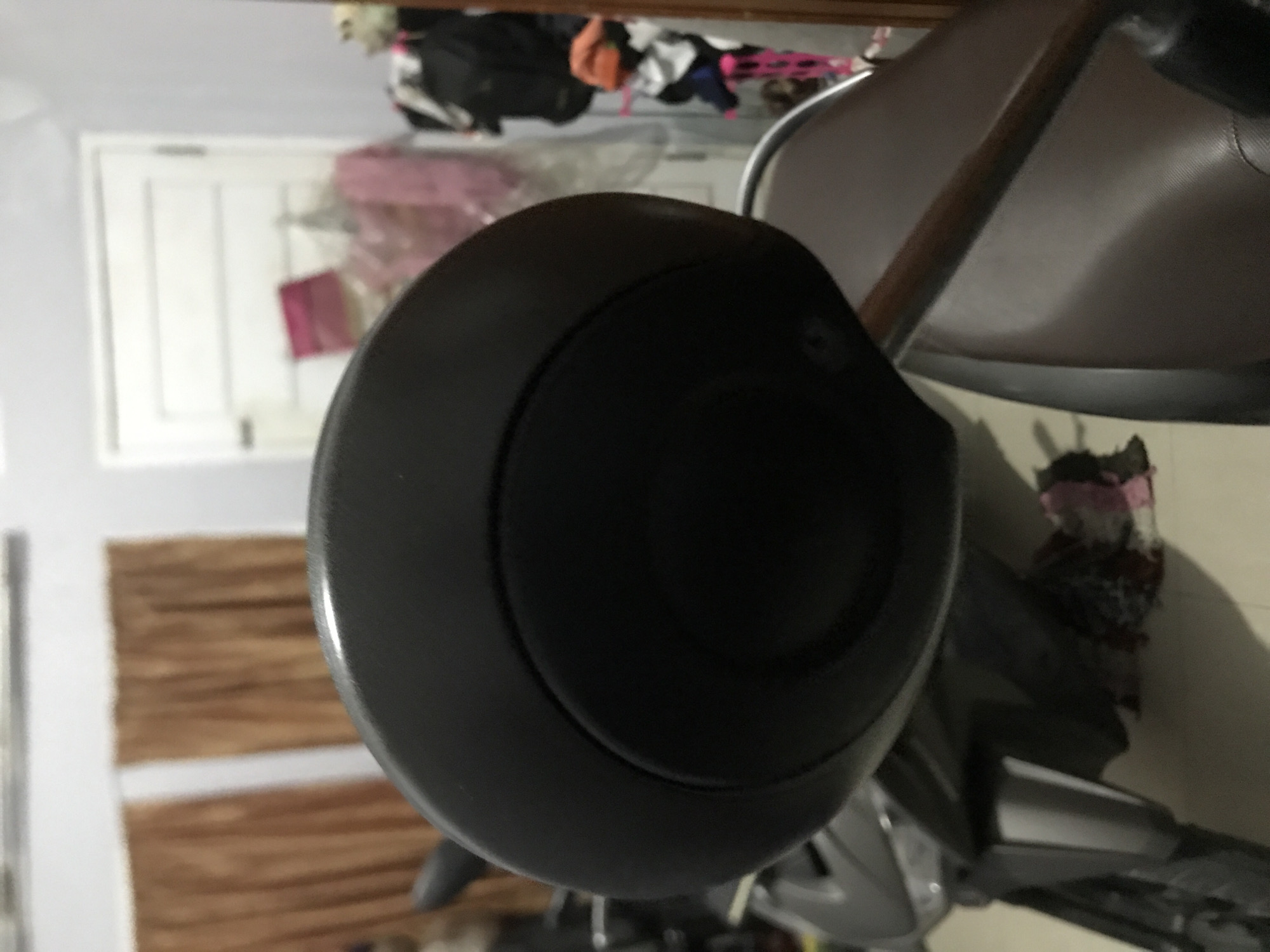 The image shows a close-up of the handlebars of a scooter, with a round, black cover over the central portion. The scooter is parked indoors, as evidenced by the interior background featuring a wooden door and miscellaneous items piled in the corner. The image does not contain enough information to determine the specific location in Lhokseumawe, Indonesia. There are no people visible.